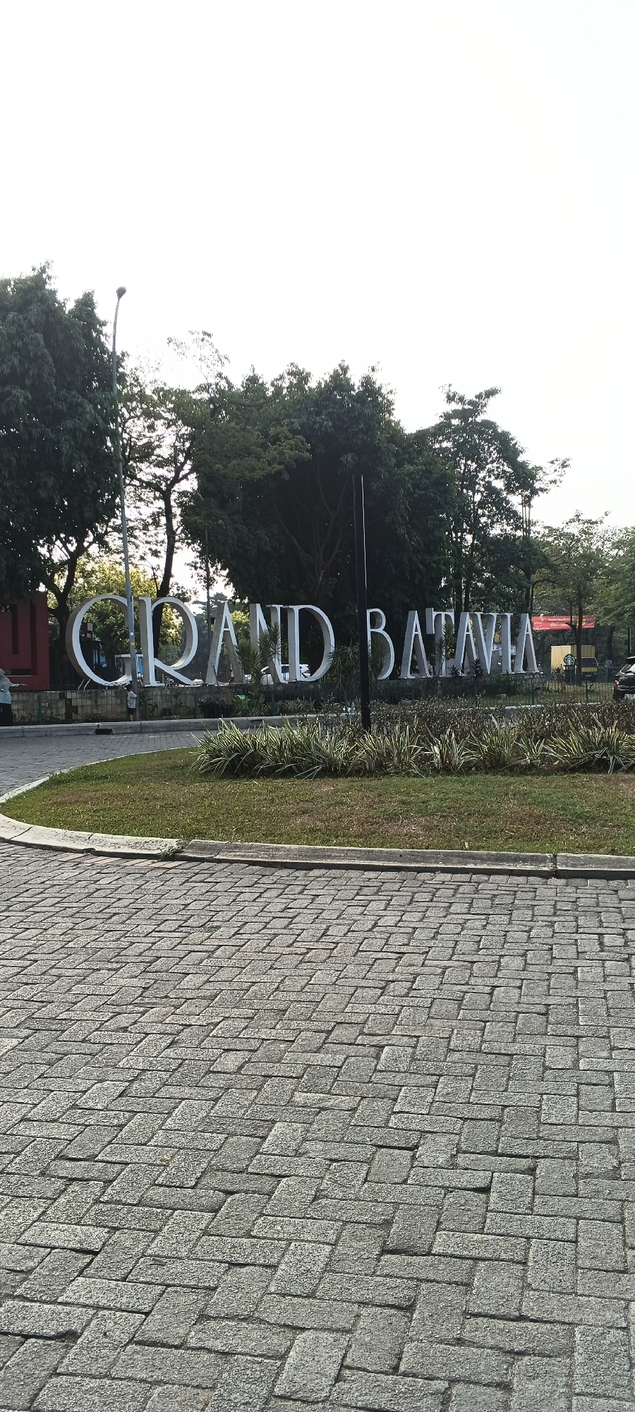 A large, three-dimensional sign spelling out "GRAND BATAVIA" is centered in the frame, set against a backdrop of trees and a white sky. The letters are white and appear to be made of a solid material, possibly concrete or metal. In the foreground, a paved area made of gray interlocking stones forms a road or walkway. To the left of the sign, a light pole stands tall, and a few people are visible near the entrance, one in a blue shirt and another in a gray shirt. Behind the sign, lush green trees and some bushes create a natural environment. Further in the background, a red structure with white lettering is visible, along with a Starbucks sign and a yellow vehicle. The overall scene suggests an entrance to a commercial area or a complex called Grand Batavia.