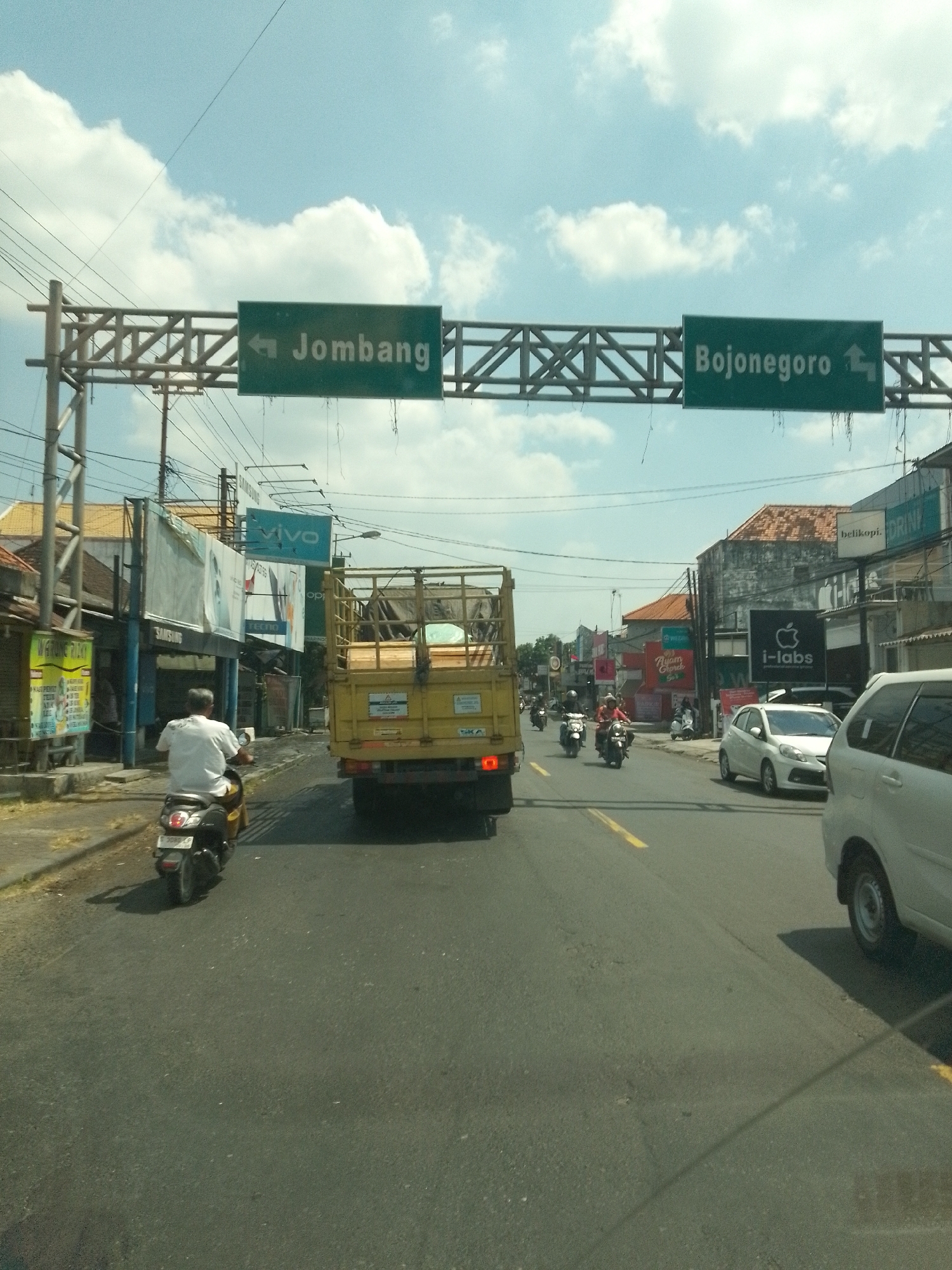 The image captures a street scene in Babat, Indonesia, under a partly cloudy sky. A highway sign with green backgrounds and white text indicates directions to "Jombang" with an arrow pointing left and "Bojonegoro" with an arrow pointing right. In the foreground, a yellow truck is moving down the road, followed by a white car on the right side. A person on a black motorcycle is visible on the left side of the road, heading away from the viewer. Further down the road, more motorcycles and a white car can be seen. The street is lined with buildings and shops, including a "VIVO" sign and other commercial advertisements. Power lines are strung overhead, and vegetation is visible along the roadside. The road surface is asphalt with yellow lane markings.