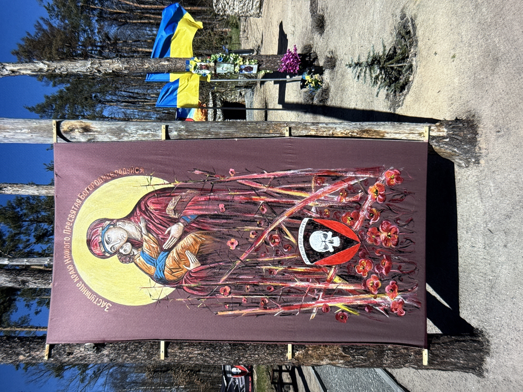 In an outdoor setting with trees and blue sky, a large banner with religious iconography is displayed. The banner, in shades of brown and red, features an image of the Virgin Mary holding the infant Jesus, rendered in a traditional iconographic style with a yellow halo. The image is surrounded by depicted red flowers and thorny branches. Below the central image, a shield with a skull insignia is visible. The banner is suspended between two rough wooden posts. In the background, two Ukrainian flags are visible, one fully unfurled and the other partially furled, attached to a tree. Additional flags and memorial items, including framed pictures and floral arrangements, are placed around the base of the tree. A small evergreen tree stands to the right of the banner. The ground is a sandy, gravelly surface.