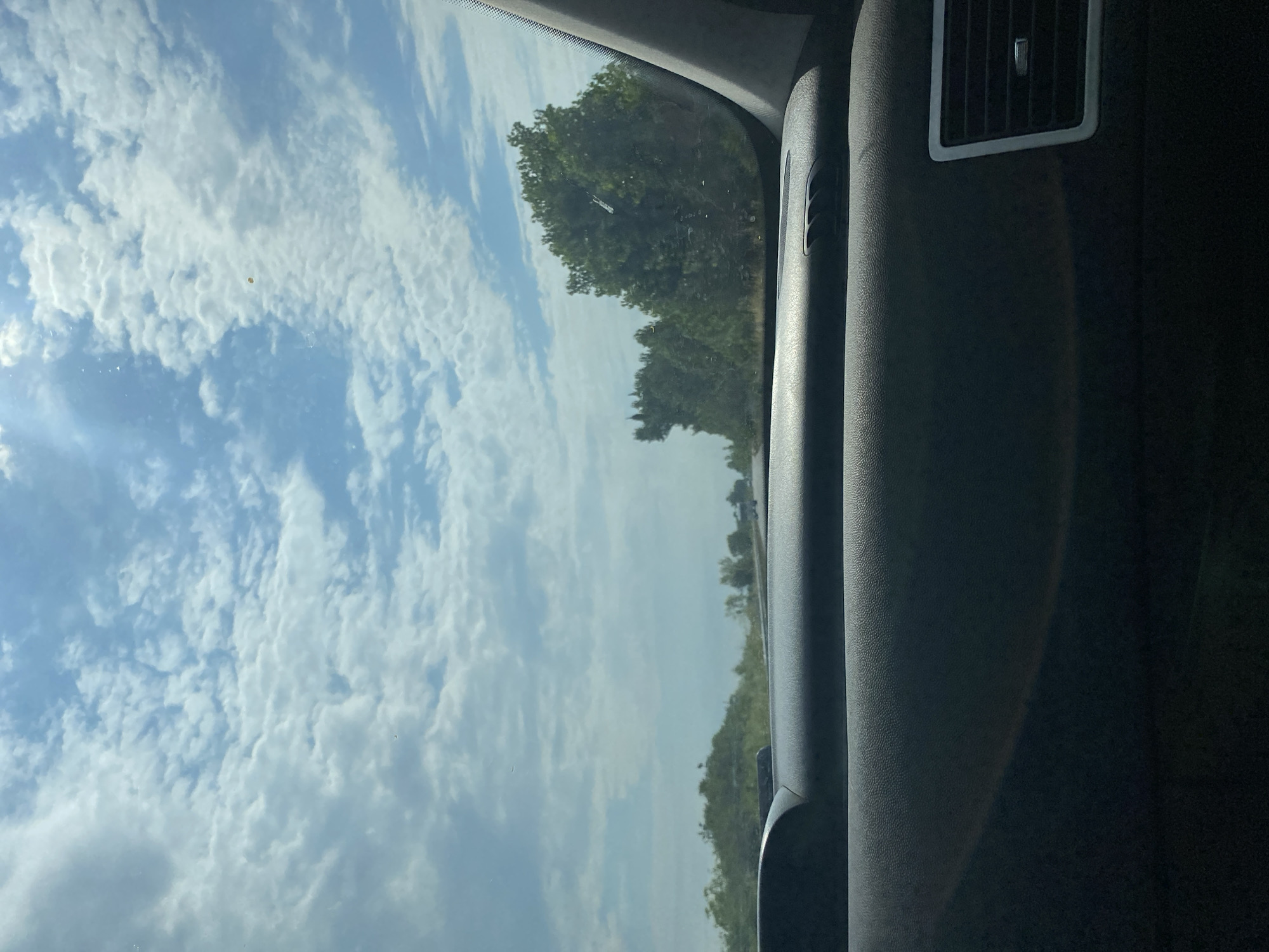 This is a vertical, medium-shot photograph taken from inside a vehicle, looking out the passenger-side window. The view captures a bright, partly cloudy sky with fluffy, white clouds scattered against a light blue backdrop. Sunlight streams through the clouds, creating a slightly hazy effect.
To the right of the frame, the interior of the car is visible, dominated by dark grey textured plastic. A silver-trimmed air vent is prominent in the upper right corner of the image. The thick pillar of the car's frame divides the interior from the exterior view.
Outside the car, a line of lush green trees forms a distant treeline against the sky. The trees suggest a rural or suburban setting, possibly during daytime given the brightness. The image gives the impression of travel, perhaps on a highway or a scenic route.
There is no visible text in the image, and no people are present. The overall mood is serene and suggests a pleasant journey on a clear, albeit cloudy, day. The crops highlight the textures of the clouds and the car's interior, as well as the detail of the air vent.
