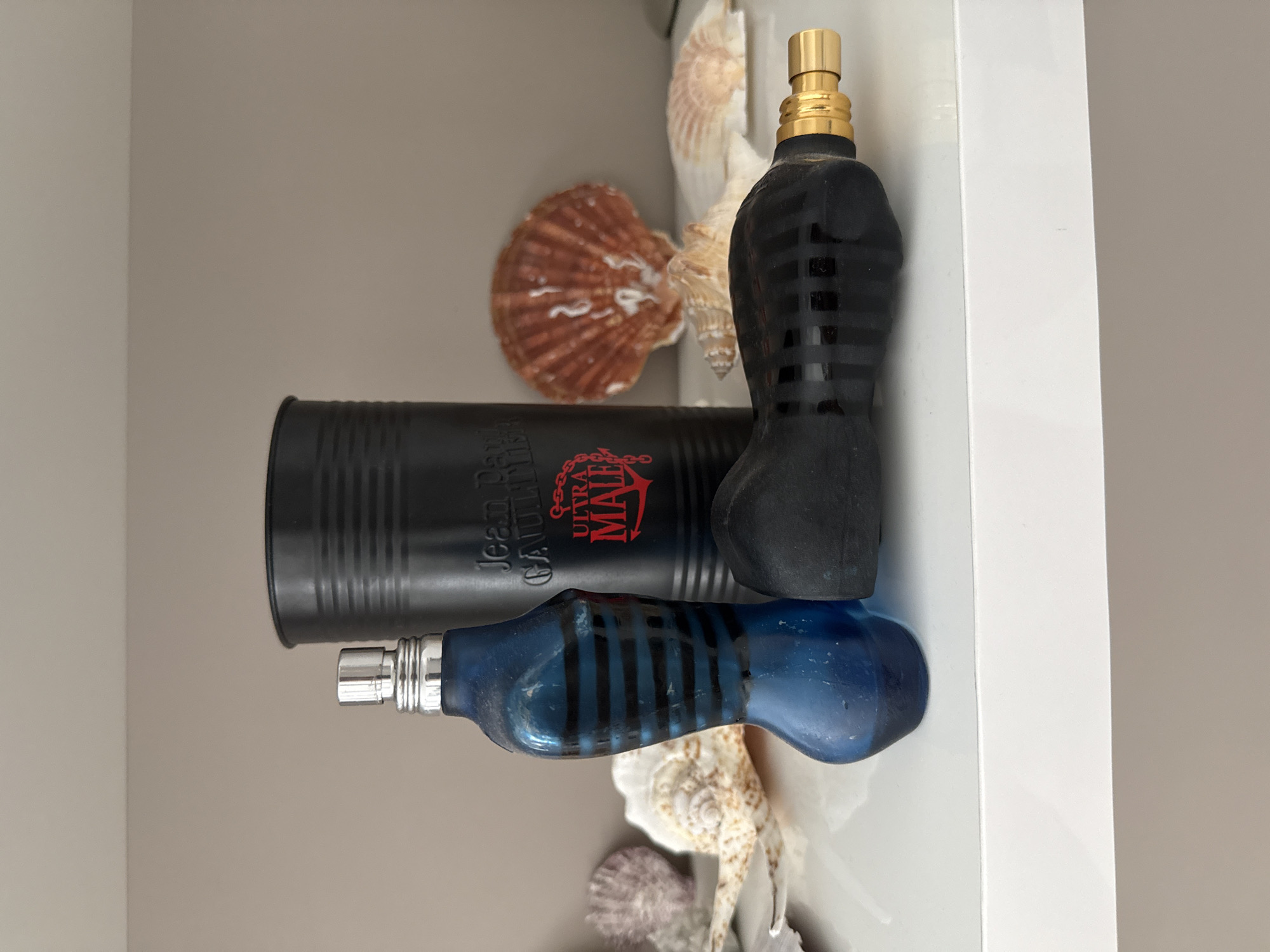 This is a still life image featuring two perfume bottles and a cylindrical container, arranged on a white shelf against a neutral, beige-toned wall. In the foreground, a blue perfume bottle, shaped like a human torso with black stripes, stands upright. To its right, a black perfume bottle, also with black stripes and a gold nozzle, lies horizontally. Behind them, a black cylindrical container with ribbed texture displays the text "Jean Paul GAULTIER ULTRA MALE" in white and red lettering. Various seashells, including a large fan-shaped one in brown and white and smaller, elongated shells, are scattered around the bottles. The lighting is soft, creating a gentle shadow cast by the objects. The scene is static and appears to be an indoor display. The location is not discernible from the image itself; it is not in Burgas, Bulgaria.