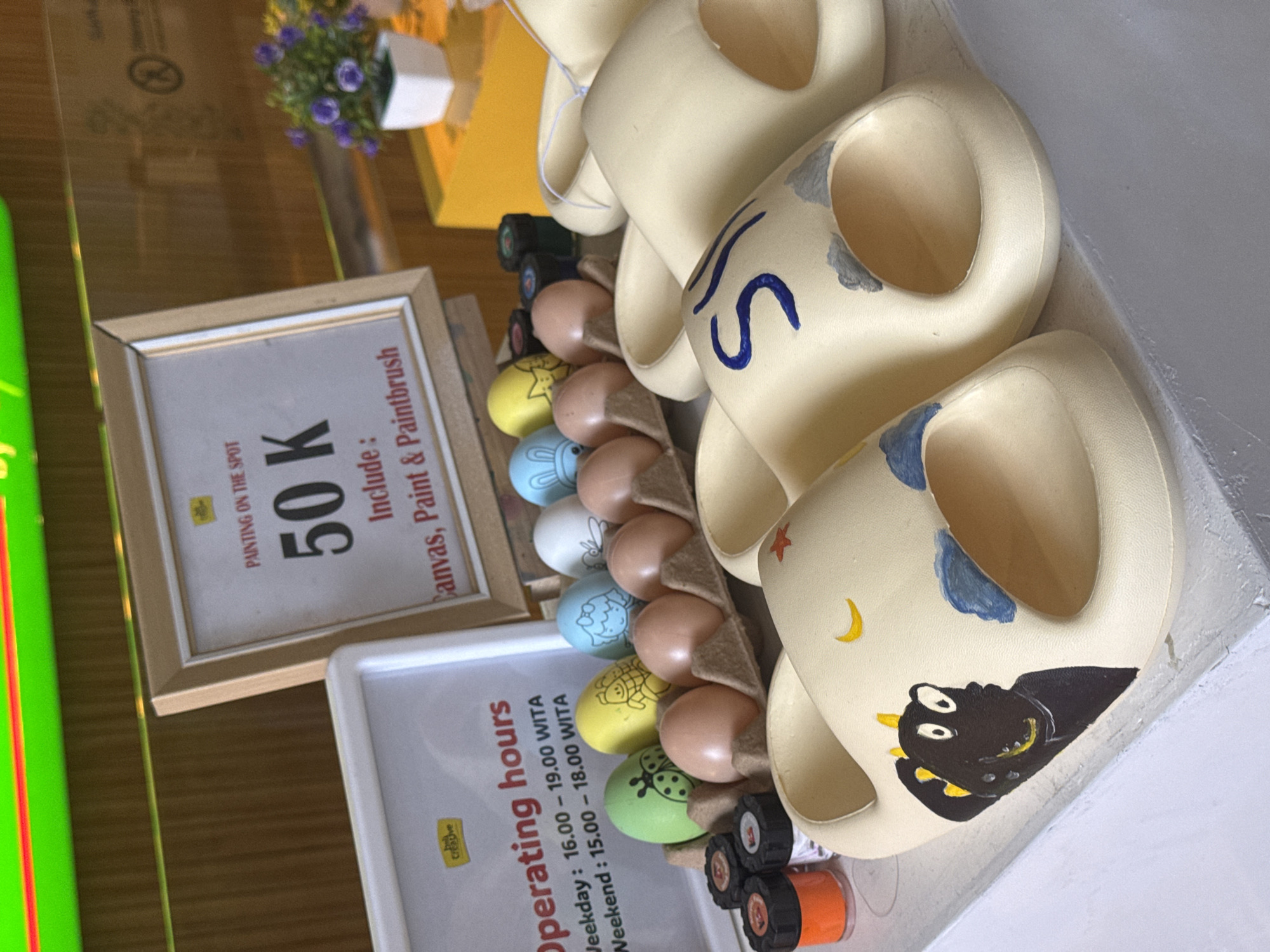 An indoor scene, rotated clockwise, displays items associated with a creative painting activity, likely in Kuta Selatan, Indonesia. The main subjects are several beige ceramic mugs or objects, some partially painted with designs like a dark creature, a crescent moon, stars, clouds, and the letter "S." Alongside these, a carton holds a mix of natural brown eggs and eggs painted with various cartoon-like figures, including a ladybug, fish, butterfly, and bunny. Small pots of paint in colors like orange, black, yellow, and blue are visible, suggesting the materials used for the artwork.
Two framed signs provide information. One sign, titled "PAINTING ON THE SPOT," advertises a price of "50 K" (likely Indonesian Rupiah) and states that it "Include: Canvas, Paint & Paintbrush." The other sign lists "Operating hours," indicating "Weekday: 16.00 – 19.00 WITA" and "Weekend: 15.00 – 18.00 WITA." "WITA" refers to Central Indonesian Time. A small pot with purple artificial flowers and a bright green illuminated strip on the left add to the setting. The arrangement suggests a craft studio or a creative corner where customers can engage in painting activities. The lighting is bright, indicating either daytime or a well-lit interior.