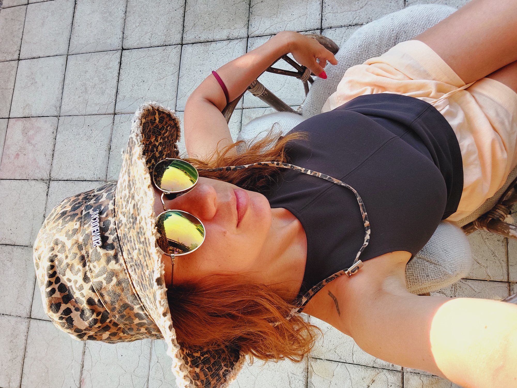 A woman is sitting on a chair outdoors, possibly on a balcony or patio, given the tiled flooring visible in the background. She is wearing a leopard-print bucket hat, mirrored sunglasses, a black tank top, and light peach-colored shorts. A leopard-print cord is visible, attached to her hat and draped around her neck. She has red hair, and a small tattoo is visible on her left shoulder. A burgundy band is on her right upper arm. The lighting suggests it is daytime, with sunlight casting shadows. The specific location in Sozopol, Bulgaria, is not determinable from the visual information provided in the image.