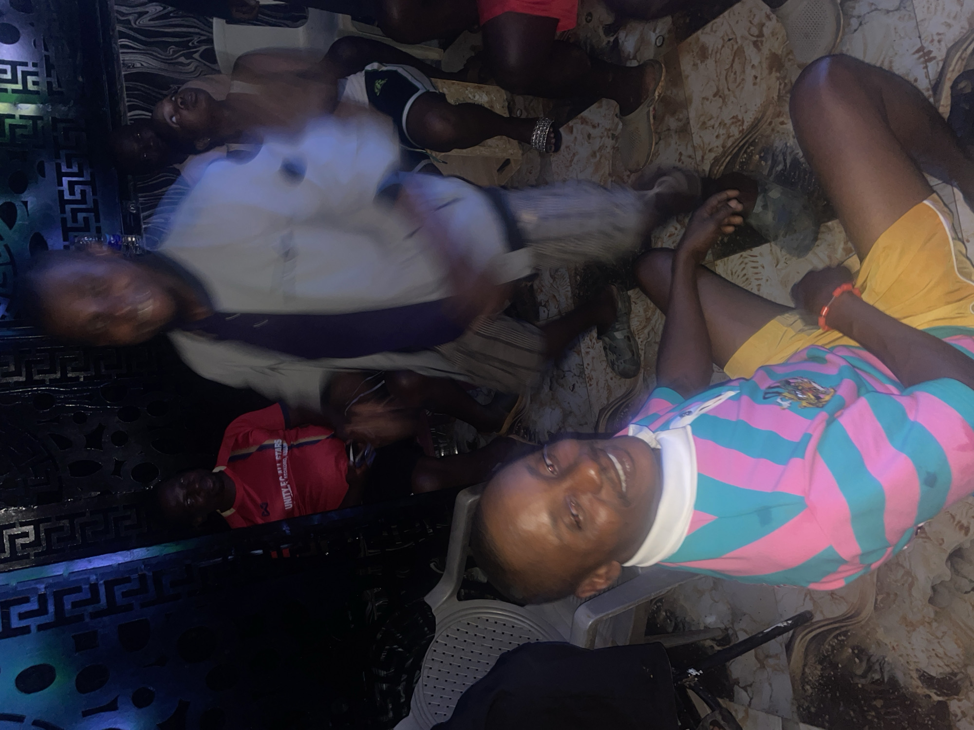 The image depicts an indoor gathering of people in what appears to be a dimly lit, possibly residential space in Kurunduma, Nigeria. The floor is tiled and appears somewhat dirty, with some debris scattered around. Several individuals are visible, mostly men, with some blurry figures suggesting movement or low light conditions.
In the foreground, a man is seated, facing the camera and smiling. He is wearing a collared shirt with alternating pink and teal stripes and a white collar. He has dark skin and short hair. To his left, another man in a red t-shirt with "UNITY FC ALL STARS" written on it is partially visible, standing and looking towards the right of the frame. Behind him, and further to the right, other individuals are present, including a woman wearing a white tank top and a man in a light-colored jacket, both of whom are blurred. The background features a dark wall with a patterned design and some vertical elements that could be part of a room divider or decorative screen. The overall impression is of a casual social gathering.