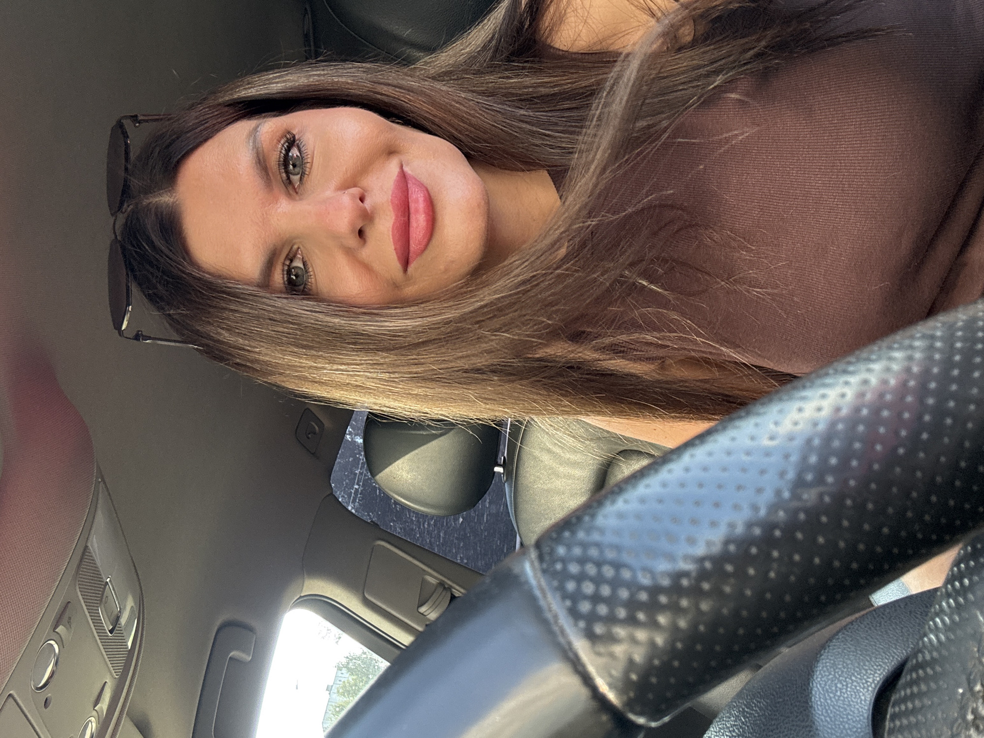 A woman with long, brown hair and sunglasses perched on her head is in the driver's seat of a car, taking a selfie. She is wearing a brown, ribbed tank top. The steering wheel of the car is visible in the foreground, and the interior of the car, including the dashboard and passenger seat, can be seen in the background. The overall lighting suggests it is daytime. No specific location in Chaikovsky, Russia, can be determined from the image.