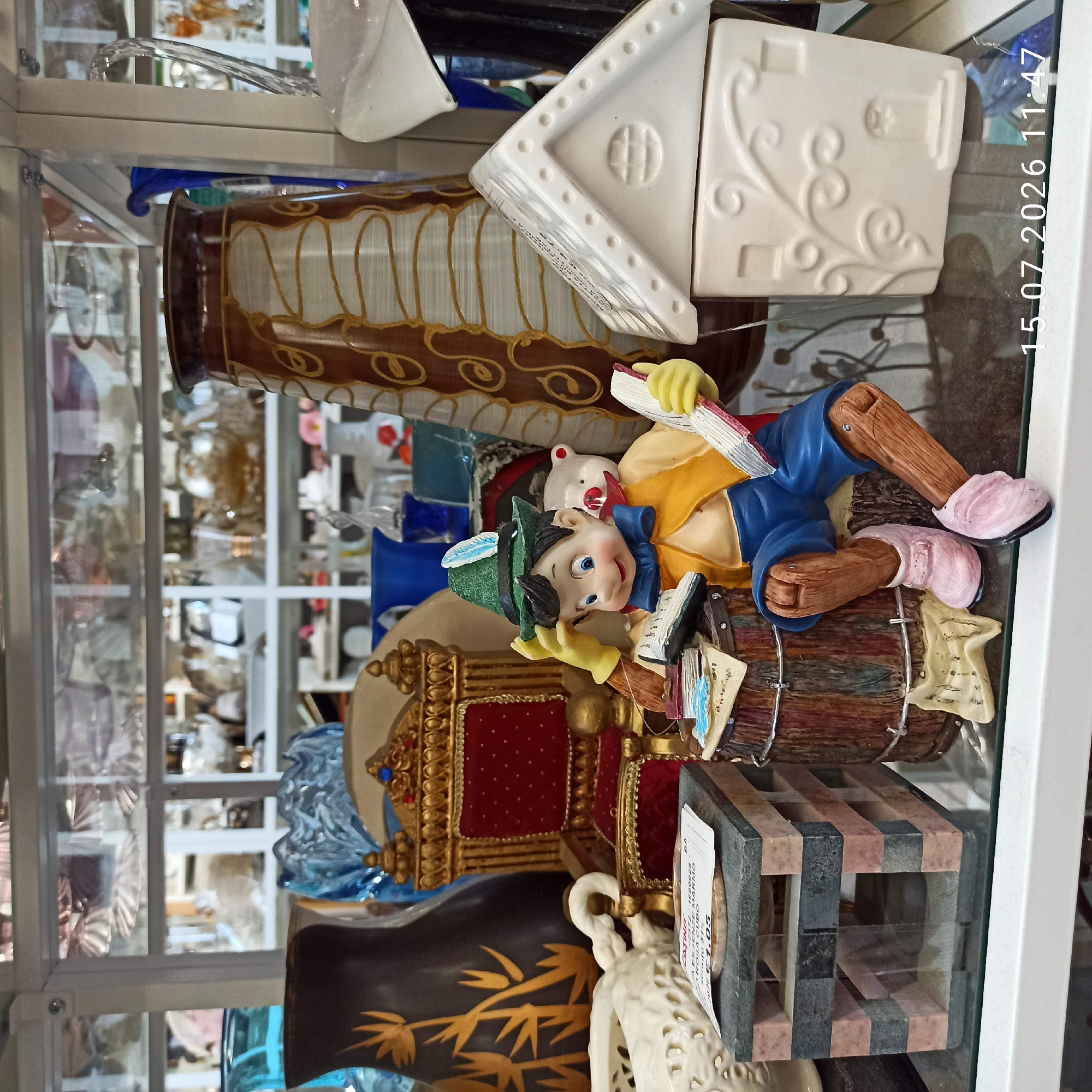 The image shows a display of various decorative items on shelves, likely in a shop. The central focus is a figurine of Pinocchio sitting on a barrel, reading a book. He is wearing a green hat with a feather, a yellow vest, and blue pants, with pink shoes. A small white bear figurine is partially visible behind Pinocchio.
To the left of Pinocchio, there is a miniature golden throne with red velvet cushions. Next to the throne is a dark brown vase with a golden bamboo-like design. A white decorative ceramic object with a patterned texture is also visible on the left. In front of the throne and vase, there is a small structure made of stacked grey and pinkish rectangular blocks, with a white label on top.
To the right of Pinocchio is a white ceramic house-shaped object with embossed floral patterns and windows. Further to the right, a large, tall vase with a brown and off-white swirling pattern is visible. In the background, shelves are filled with various glassware, vases, and other decorative items.
A timestamp "15.07.2026 11:47" is overlaid on the bottom right of the image.
The location is identified as Molfetta, Italy, implying this is a shop in that city. The scene is a still life arrangement of ornamental objects.