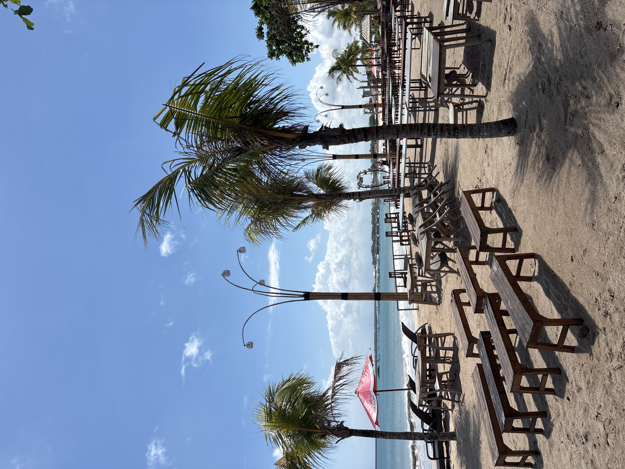 The image captures a beach scene in Kuta, Indonesia. The foreground features rows of wooden tables and benches set up on the sand, indicating a dining or event area. Several palm trees are interspersed among the tables, providing shade. In the midground, the ocean stretches out to the horizon, with a few boats visible in the distance. A red and white umbrella is planted in the sand near one of the palm trees. The sky is blue with scattered clouds. A unique, branched lamp post is positioned among the palm trees. The overall impression is a tranquil beach setting prepared for visitors.