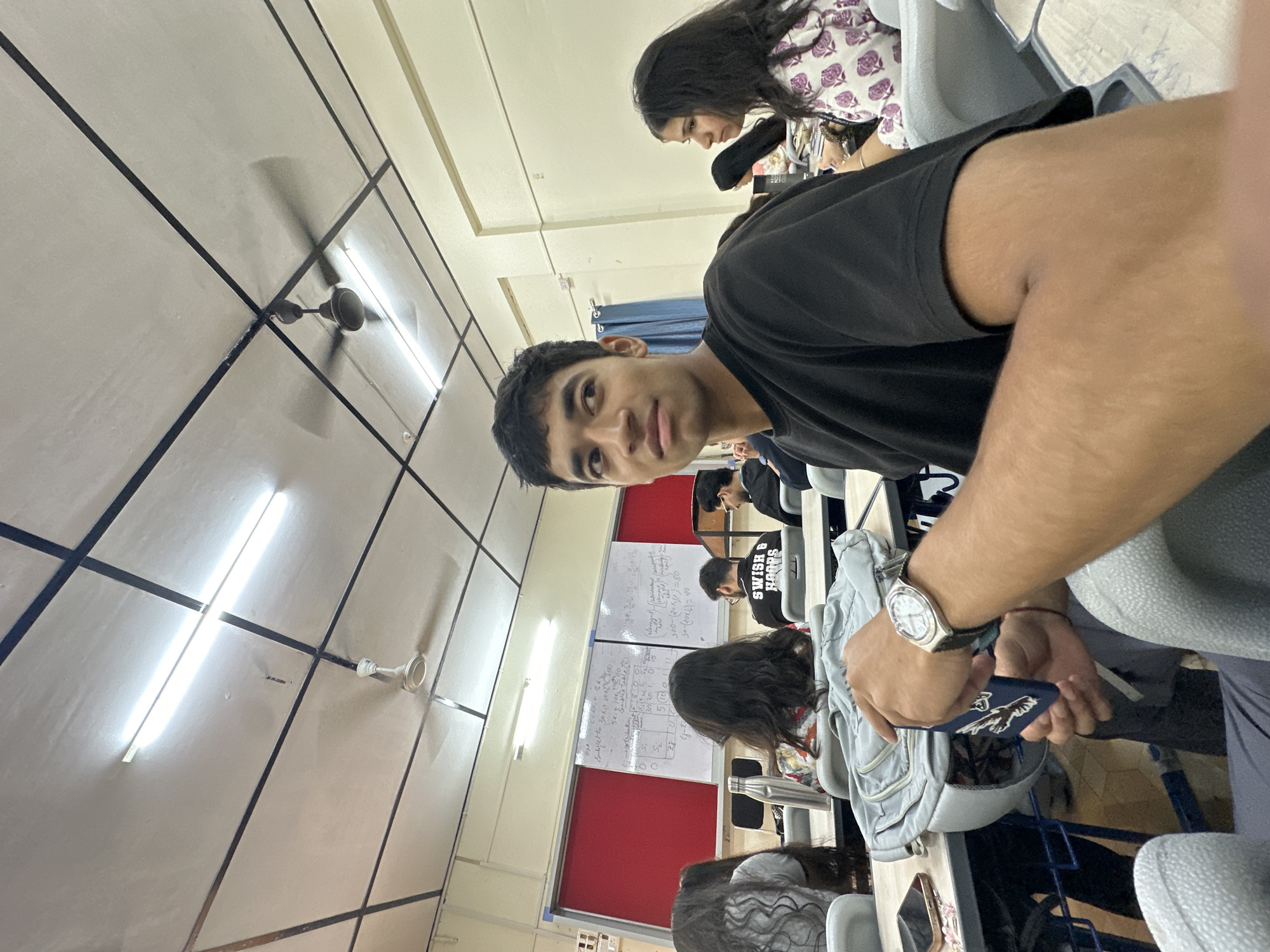 The image shows a classroom environment with students seated at desks. In the foreground, a young man wearing a black t-shirt is looking to his right, holding a dark blue object in his hands. He has a watch on his left wrist. Behind him, other students are visible, some looking towards a whiteboard with writing on it. The classroom has a tiled ceiling with fluorescent lights and ceiling fans. The overall impression is of a typical academic setting.