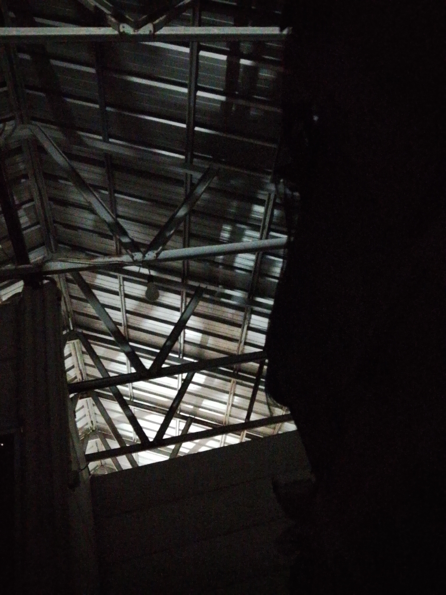 The image depicts the interior of a building in Bukit Ketok, Indonesia. The structure has a metal roof with exposed beams and a visible ceiling. The lighting is dim, and a large dark area obscures the right side of the frame. A single lightbulb hangs from the roof structure. A pillar is visible in the lower left corner.
