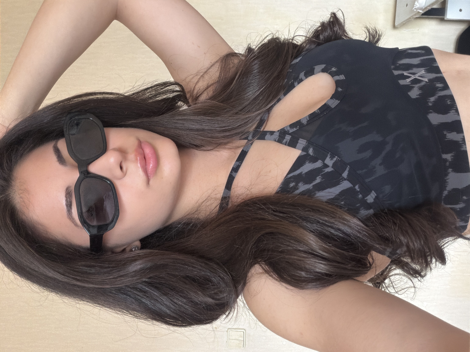 A portrait-style shot of a young woman taken from above as she lies on a light-colored surface, possibly a floor or bed, with her head oriented towards the left of the frame. Her dark, long wavy hair spreads out around her head and shoulders. She is wearing black rectangular sunglasses and has a hint of pink lip gloss on her lips. She is dressed in a dark-colored sports bra or crop top with a gray animal print pattern and criss-cross straps visible on the front. One arm is raised above her head, partially visible. The background is a plain, light-colored surface, and there's a small white electrical outlet visible towards the bottom right of the image. In the upper right corner, a piece of white furniture or fixture can be vaguely seen. The overall lighting suggests an indoor setting, possibly during the daytime. No specific location cues to Sofia, Bulgaria are visible in the image itself.