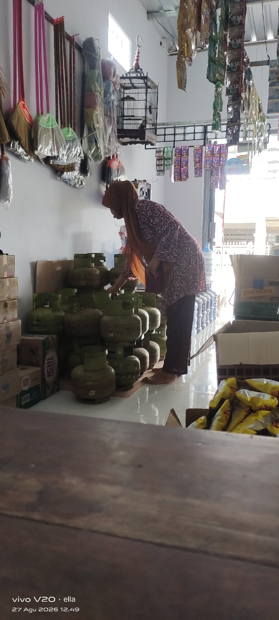 In Simo, Indonesia, a woman wearing a brown hijab and a patterned maroon and white blouse is seen tending to a stack of green gas cylinders in what appears to be a small shop or warehouse. She is bending down, her right hand extended as if to adjust or pick up one of the cylinders. The gas cylinders are arranged in multiple rows, some stacked vertically. To the left of the cylinders, several cardboard boxes are stacked, with labels visible including "ICHI OCHA". In the background, various items are hanging from the ceiling, including what look like brooms, bags of products, and a birdcage. Further back, a large water cooler can be seen. On the right side of the image, a cardboard box with "ENERGEN" printed on it is visible, along with bags of yellow snacks. The flooring is tiled. The overall impression is of a retail or storage space stocking household goods and food items.