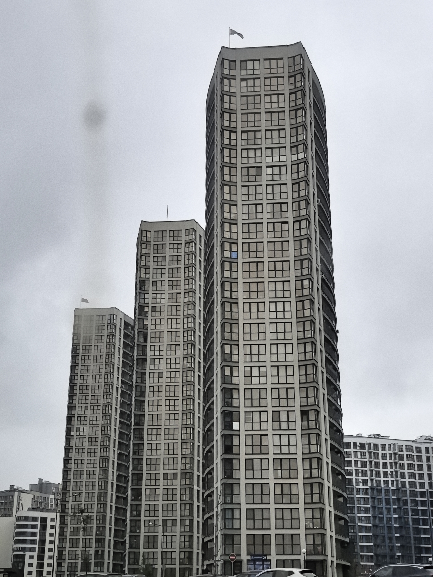 An exterior, low-angle, full shot captures a cluster of modern, high-rise apartment buildings under a cloudy, overcast sky. The buildings are predominantly gray and white, with numerous windows. The tallest building, located on the right, features a slightly curved facade and a flag at its apex. To its left, two other similar buildings are visible, also with many windows. In the background, a longer, lower building with a different architectural style, featuring a checkered pattern of windows, is also present. A dark, circular smudge is visible in the upper left quadrant of the sky, likely a lens artifact. The overall impression is of a contemporary urban residential development.
