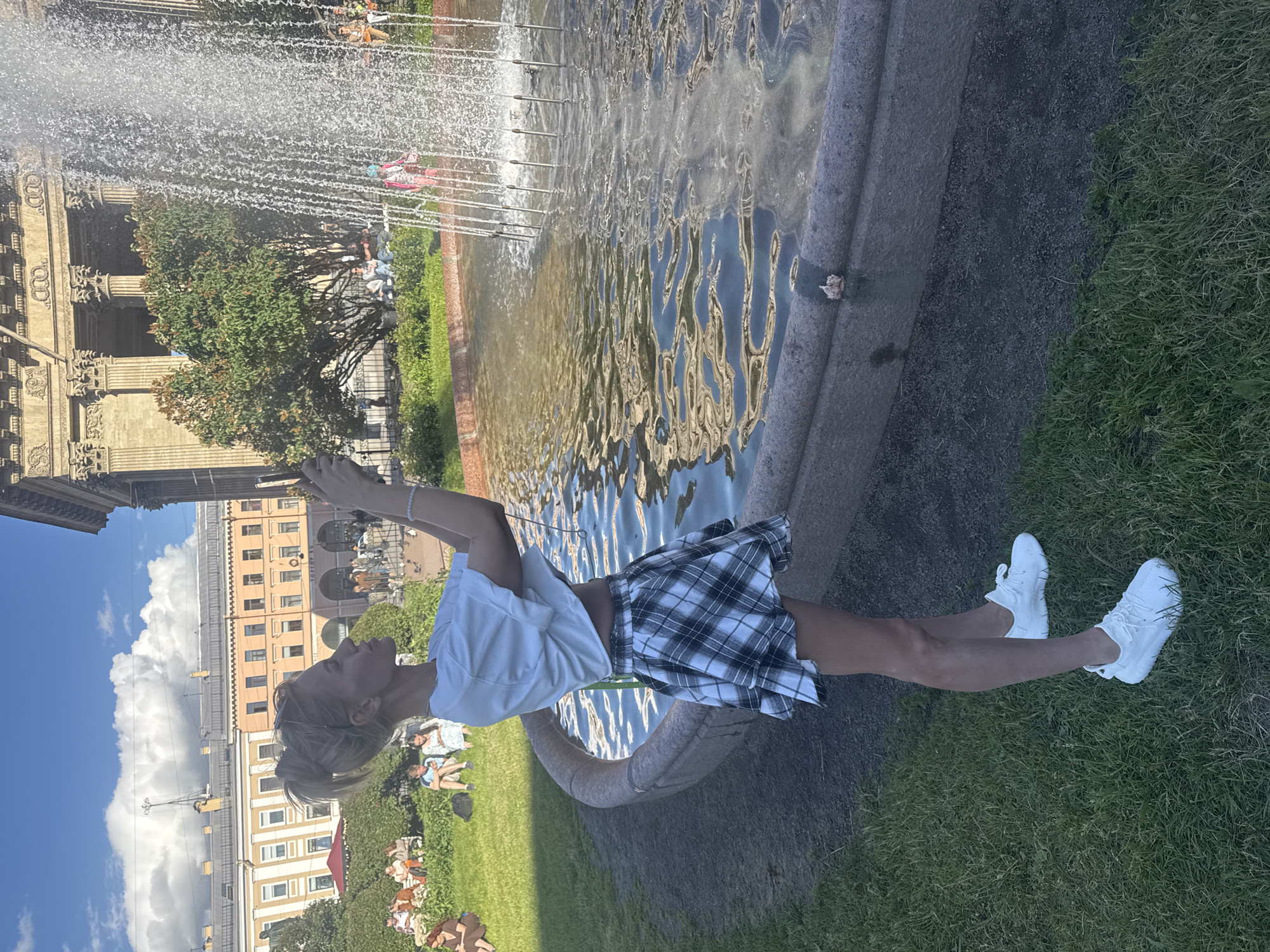 A woman stands in front of a fountain in Saint Petersburg, Russia, holding a phone to take a picture. She is wearing a white crop top and a black and white plaid skirt, with white sneakers. The fountain is in the foreground, with water spraying upwards. In the background, there are people sitting on benches on a grassy lawn, and a large building with columns and a classical facade. The sky is blue with white clouds. The location is likely Kazan Cathedral in Saint Petersburg.