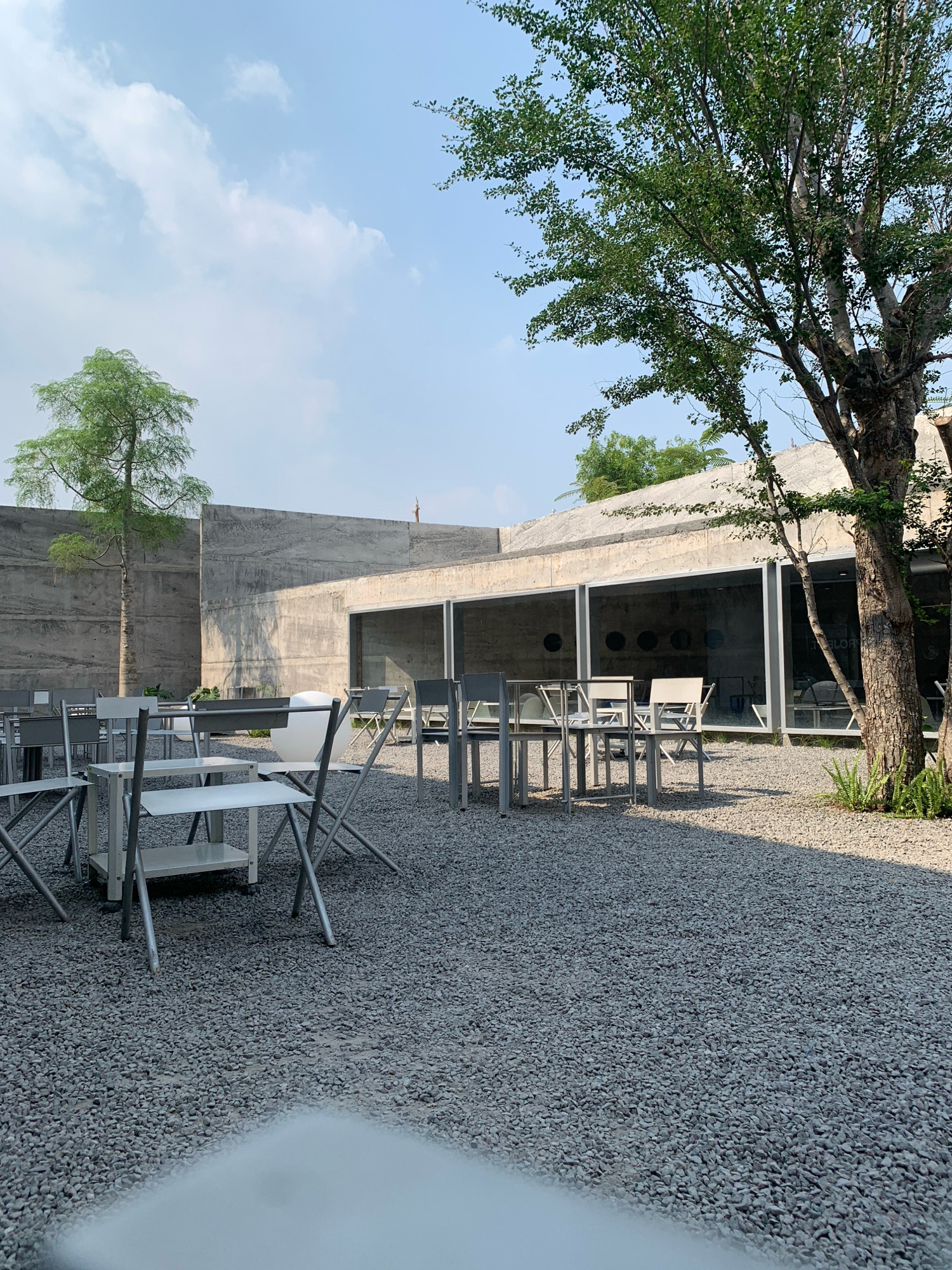 The image depicts an outdoor seating area with minimalist concrete architecture and gravel ground. Several tables and chairs are arranged in the gravel courtyard, suggesting a cafe or restaurant setting. A large tree with green foliage stands to the right, casting a shadow on the ground. To the left, a slender tree is visible in front of a tall, smooth concrete wall. The background features a modern building with large glass windows, revealing a glimpse of the interior. The sky is partly cloudy with a clear blue expanse. The scene appears to be in a serene, possibly urban, environment in Meri, Indonesia. There are no people visible in the image.