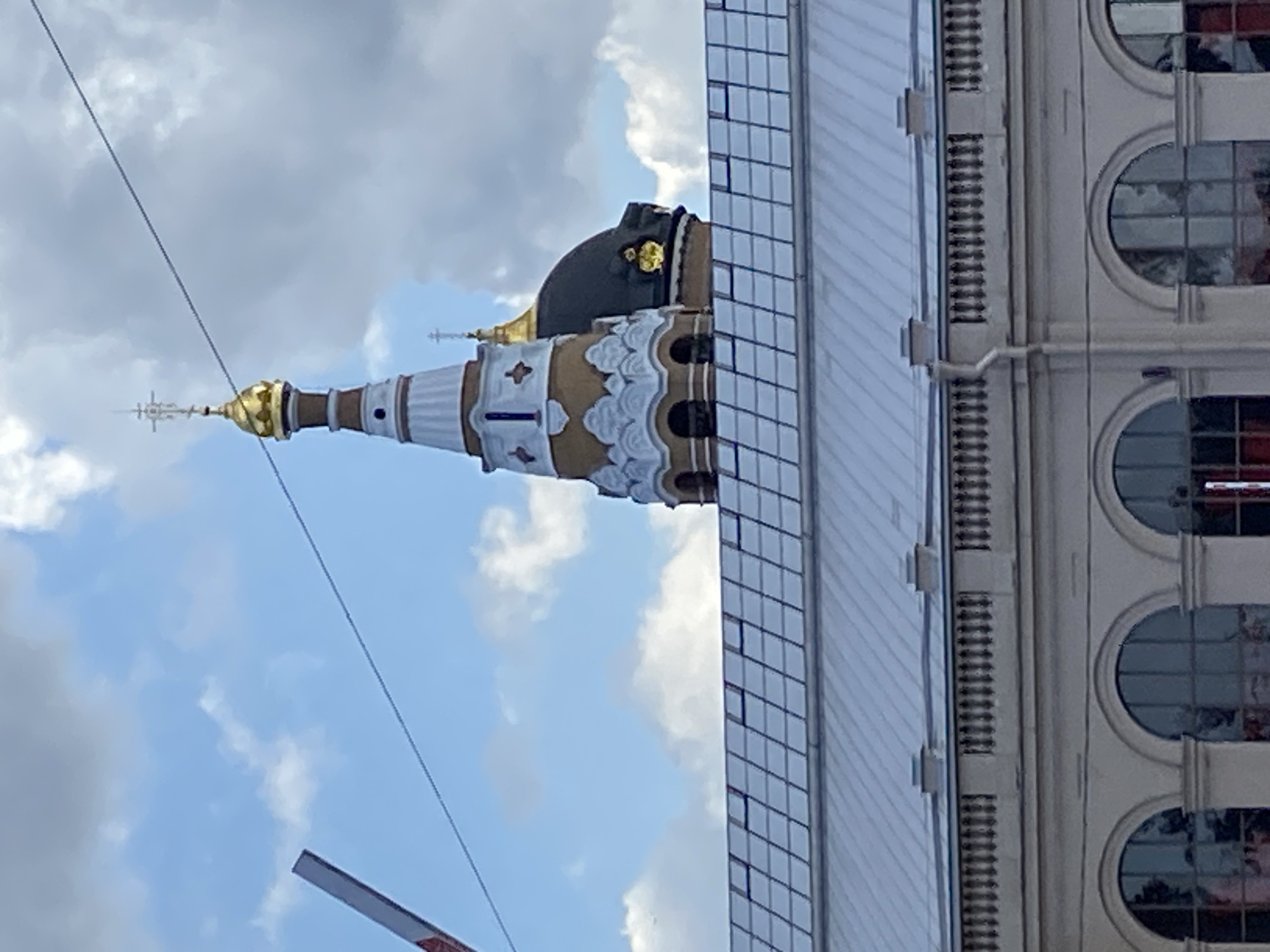 The image depicts a view of the city of Saint Petersburg, Russia, focusing on the upper portions of a building and a church tower. In the foreground, a multi-story building with arched windows and a decorative balustrade is visible. Above this building, a large, slanted roof with a grid pattern is prominent. The background features a sky with scattered clouds, and a tall, slender church tower with a golden dome and a cross at its peak. A smaller dark dome, likely from another part of the church or a nearby structure, is also visible. A thin wire runs diagonally across the sky. A red and white striped object is partially visible in the upper left corner of the frame, suggesting the presence of a barrier or some other form of temporary structure. The overall scene is dominated by architectural elements and the sky, with no people clearly discernible.