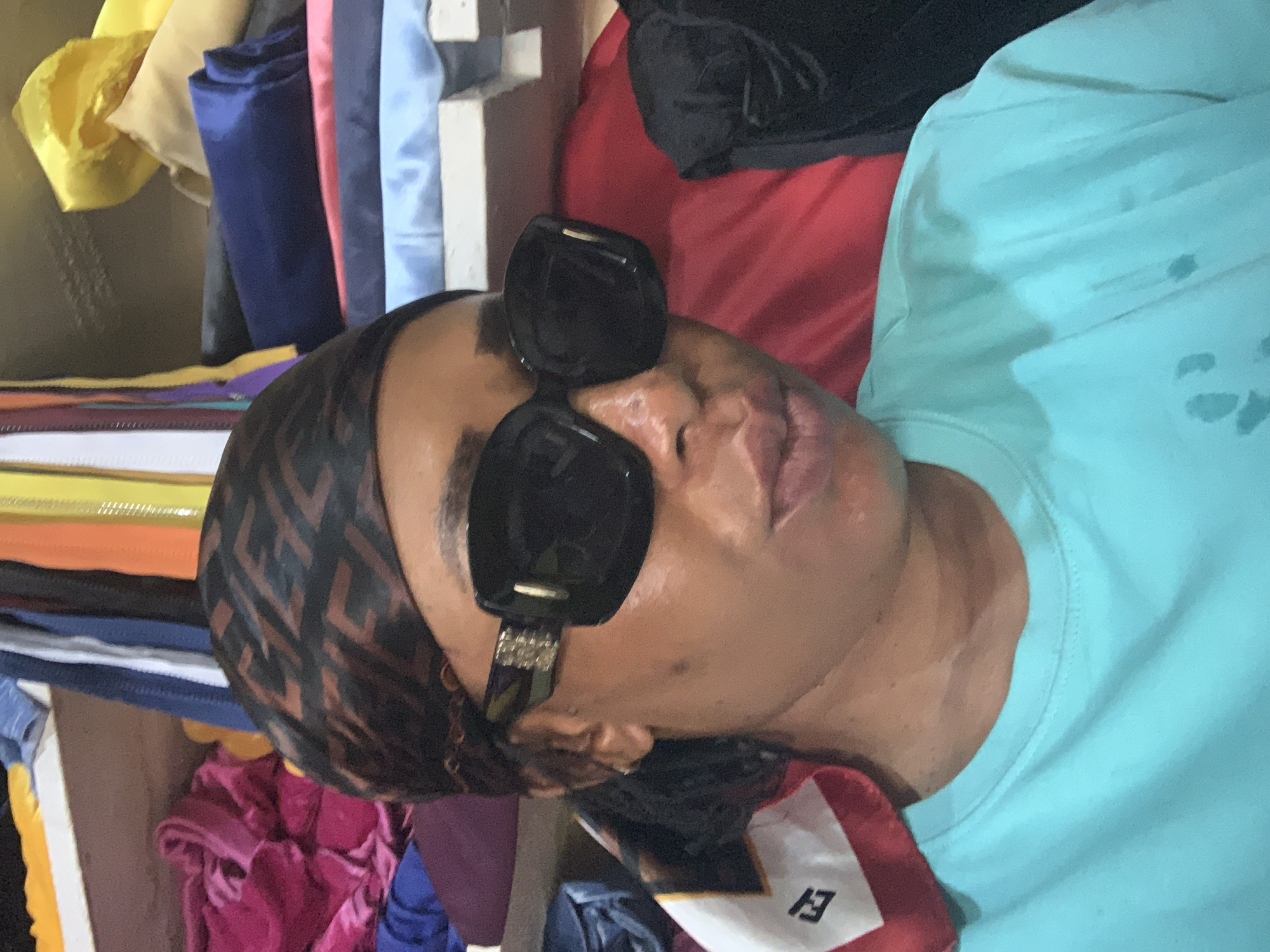 The image shows a person wearing sunglasses and a designer head covering, lying down with a light blue t-shirt visible. They appear to be in a shop or storage area with rolls of fabric in various colors stacked around them. The surroundings suggest a commercial environment, possibly a textile or clothing store in Sabon Tasha, Nigeria.