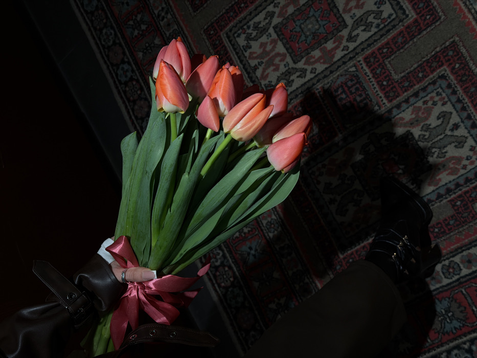An overhead, angled shot depicts a person's lower body and arm holding a large bouquet of tulips in a dimly lit indoor setting in Yerevan, Armenia. The prominent subject is a bouquet consisting of approximately 10-12 coral and orange-pink tulips with long green stems and leaves, tied with a wide, dark pink satin ribbon.
The person's left hand is visible, featuring manicured nails with white tips and a silver-colored ring on the ring finger, gripping the bouquet. The wrist is adorned by a dark brown leather-like jacket sleeve with a buckle and strap detail, under which a white cuff is visible. The lower left portion of the image shows a dark-clad leg wearing a dark boot with a heel and multiple decorative straps/buckles.
The floor is covered by an ornate, traditional rug featuring geometric and stylized patterns in shades of red, dark blue, beige, and green. The scene is characterized by dramatic lighting, with a strong light source illuminating the bouquet and portions of the rug, casting defined shadows.