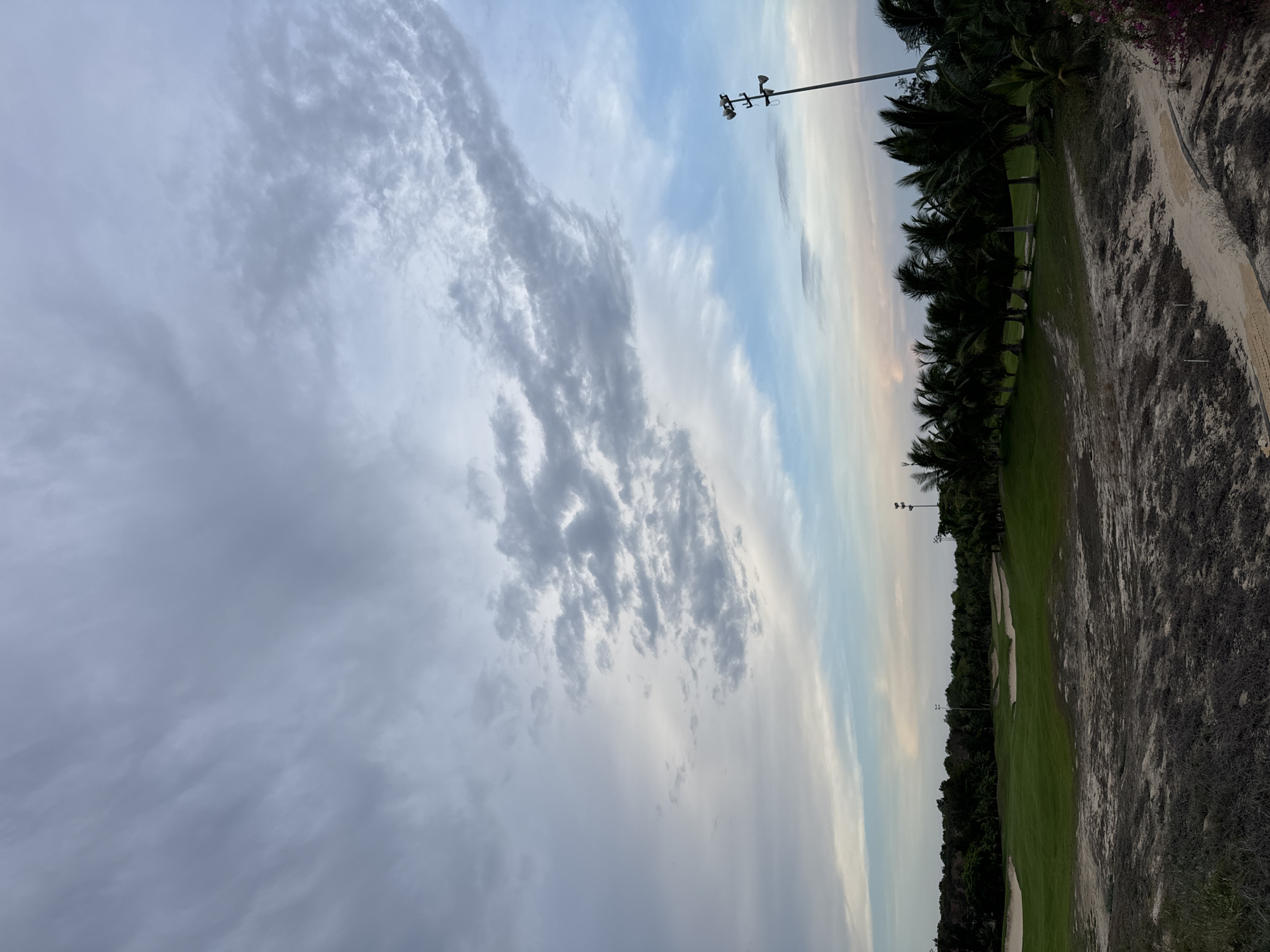 The image depicts a golf course in Mui Ne, Vietnam, under a partly cloudy sky. To the right is a strip of sand, possibly a walking path or rough area, and it appears a row of palm trees lines the edge of the course. The golf course itself is green, interrupted by sand traps. A light pole with some kind of electrical or weather equipment is visible on the right. The clouds are predominantly gray, with some blue peeking through. The image is oriented vertically.