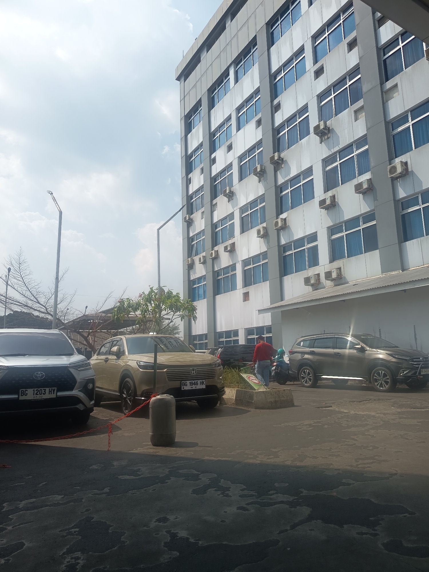 The image captures a daytime scene outside a modern, multi-story building with a facade of grey panels and numerous blue-tinted windows. Several air conditioning units are visible mounted on the exterior walls. In the foreground, the paved ground shows a pattern of light and dark patches, suggesting it might be wet or stained.
Parked on the paved area are several vehicles: a silver SUV on the left, a beige SUV in the middle, and a dark grey SUV on the right. The silver SUV has a license plate that reads "BG 1203 HY". The beige SUV has a license plate "BG 1946 HK". The dark grey SUV's license plate is partially visible, showing "BG 1421". A red chain is strung between the silver and beige SUVs, with a cylindrical concrete barrier between them.
A person wearing a red shirt and blue jeans is standing on the paved area between the beige and dark grey SUVs, interacting with what appears to be a motorcycle with a green helmet. The environment includes bare tree branches on the left and a small tree with some foliage behind the beige SUV. Two tall streetlights with lamps are also present, one on the far left and another behind the beige SUV. The sky above is cloudy, with patches of blue visible. The scene is consistent with an outdoor parking area or roadside in a city like Lubuklinggau, Indonesia, given the style of the building and the vehicle license plates.