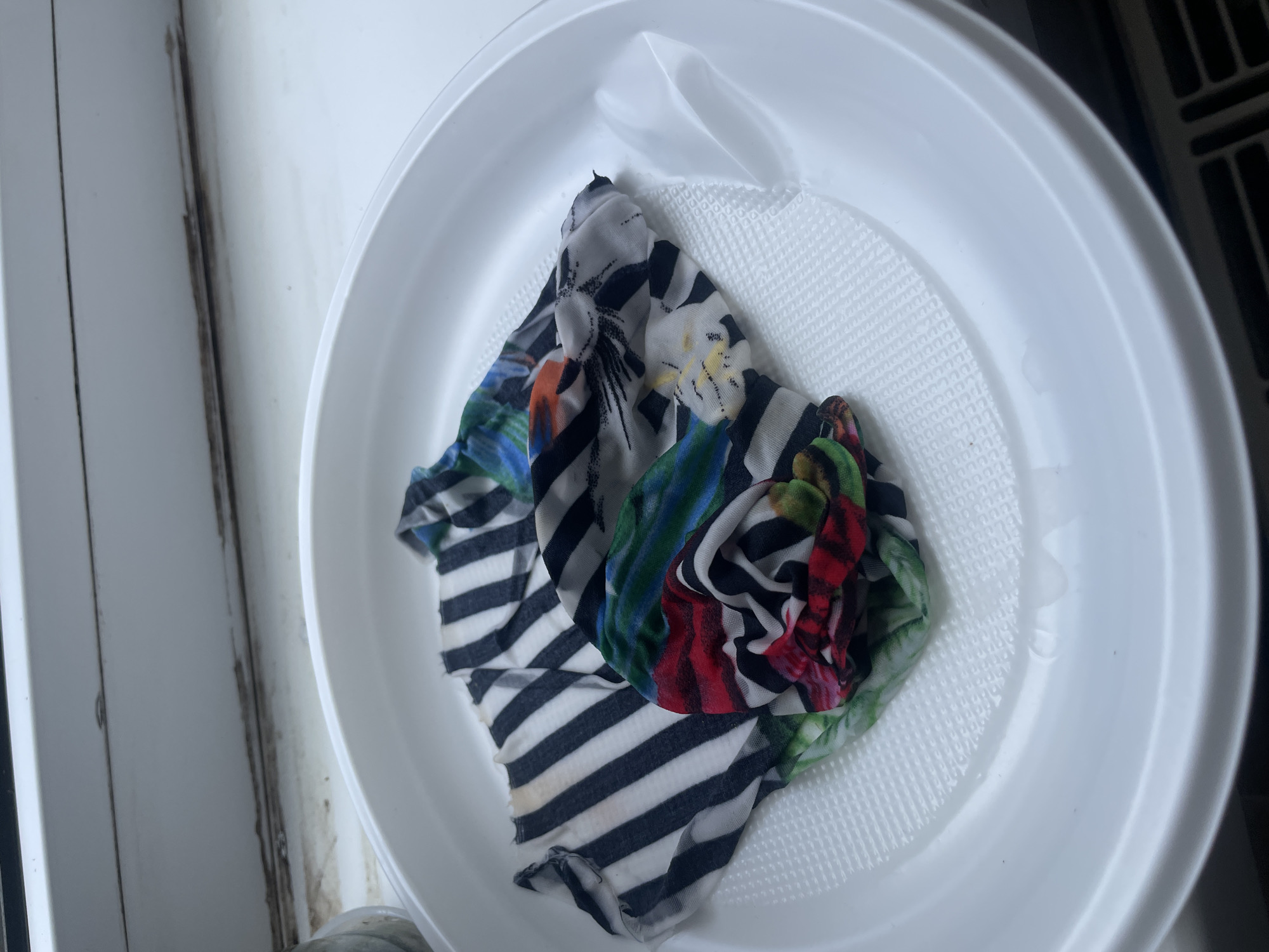 A high-angle close-up shot shows a white plastic dish filled with water. A piece of crumpled fabric with a black and white striped pattern and floral designs lies in the water. The dish is on a white surface next to a white wall. The environment appears to be indoors.