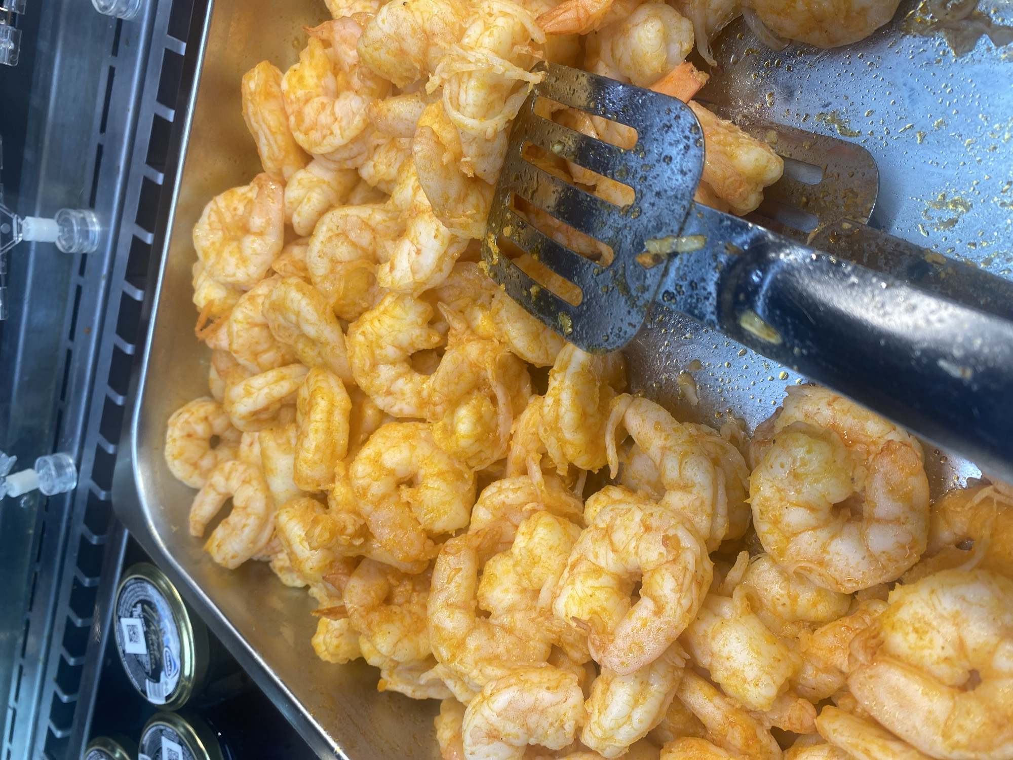 A close-up, overhead shot shows a metal tray filled with a large quantity of cooked shrimp seasoned with a reddish-orange spice mix. A portion of a metal slotted serving utensil is visible in the foreground, partially submerged in the shrimp. The shrimp appear to be cooked and ready to serve, likely in a buffet or food service setting. In the background, out of focus, are glass jars with dark contents, possibly caviar, and a metal grate with a black surface. The overall scene suggests a food display. No specific location in Khanty-Mansiysk, Russia can be determined from the image.