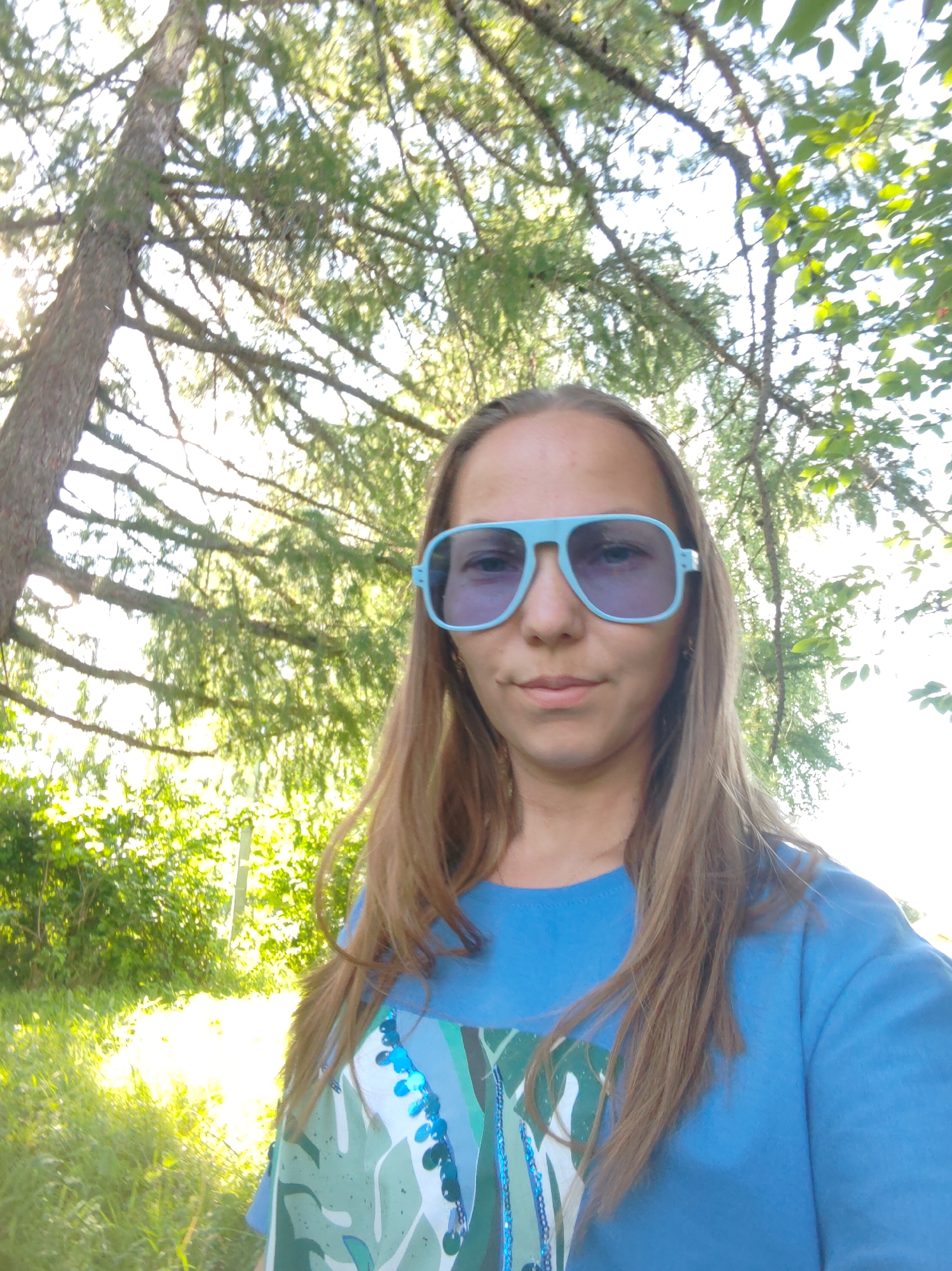 In this selfie, a young woman with long, light brown hair stands outdoors, facing the camera. She is wearing a bright blue crew-neck t-shirt with a colorful, abstract design on the front, featuring shades of green, white, and blue, accented with sequins. She has on large, light blue aviator sunglasses. The background consists of lush green foliage and trees, suggesting a park or natural setting. The sunlight filters through the leaves, creating a bright and airy atmosphere. The image appears to have been taken in the city of Chukhlem, Russia, likely during daytime.