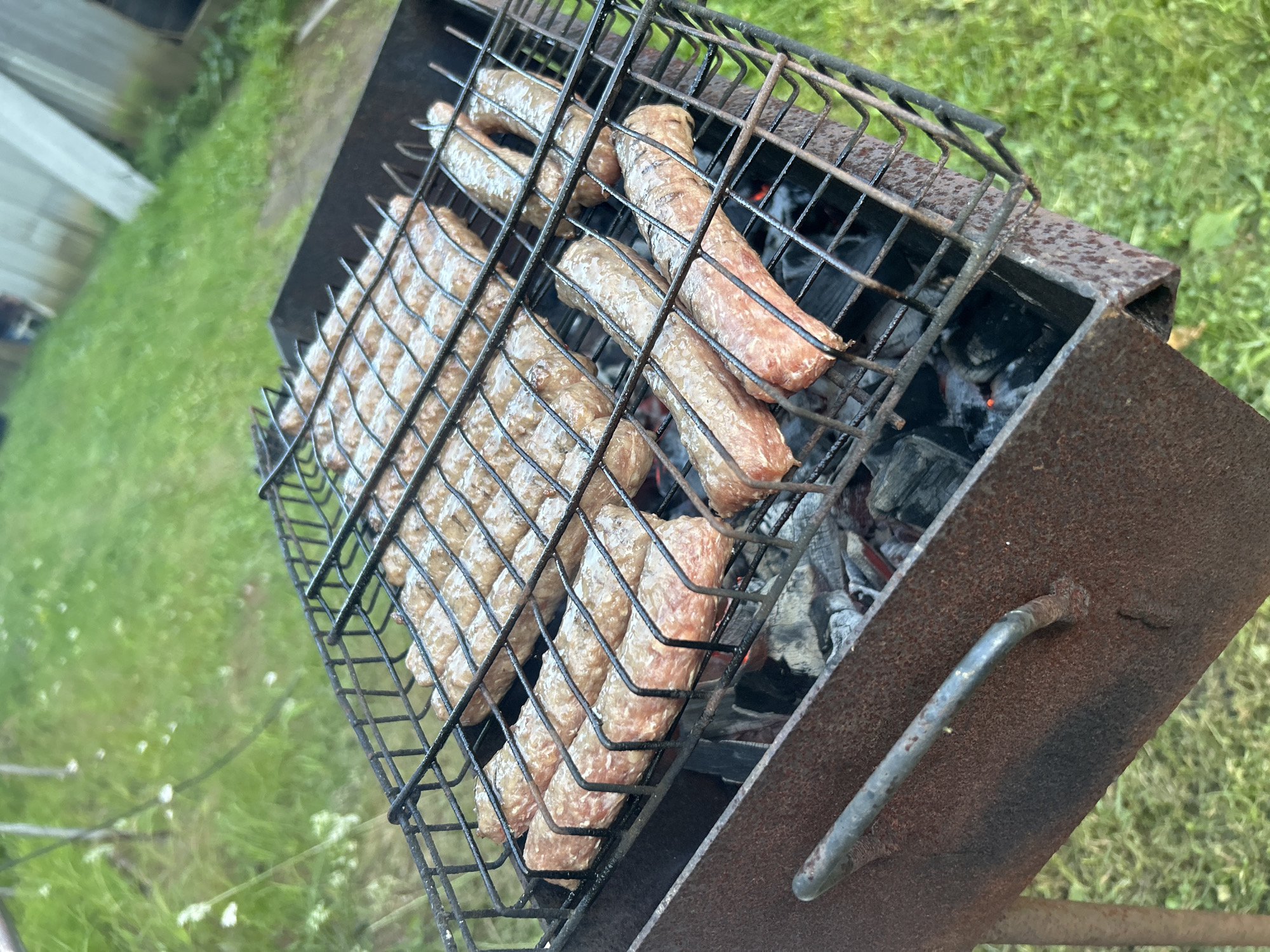 The image shows a close-up view of raw sausages on a barbecue grill. The sausages are arranged in rows on a black metal grate, which is positioned over a bed of glowing coals. The grill itself appears to be made of rusty metal, with a worn handle visible on the side. The background is out of focus, but it appears to be a grassy area with some greenery, suggesting an outdoor setting. There is no indication of people or specific urban landmarks that would allow for identification of the location in Кадряково, Russia. The scene depicts food preparation in an outdoor environment.