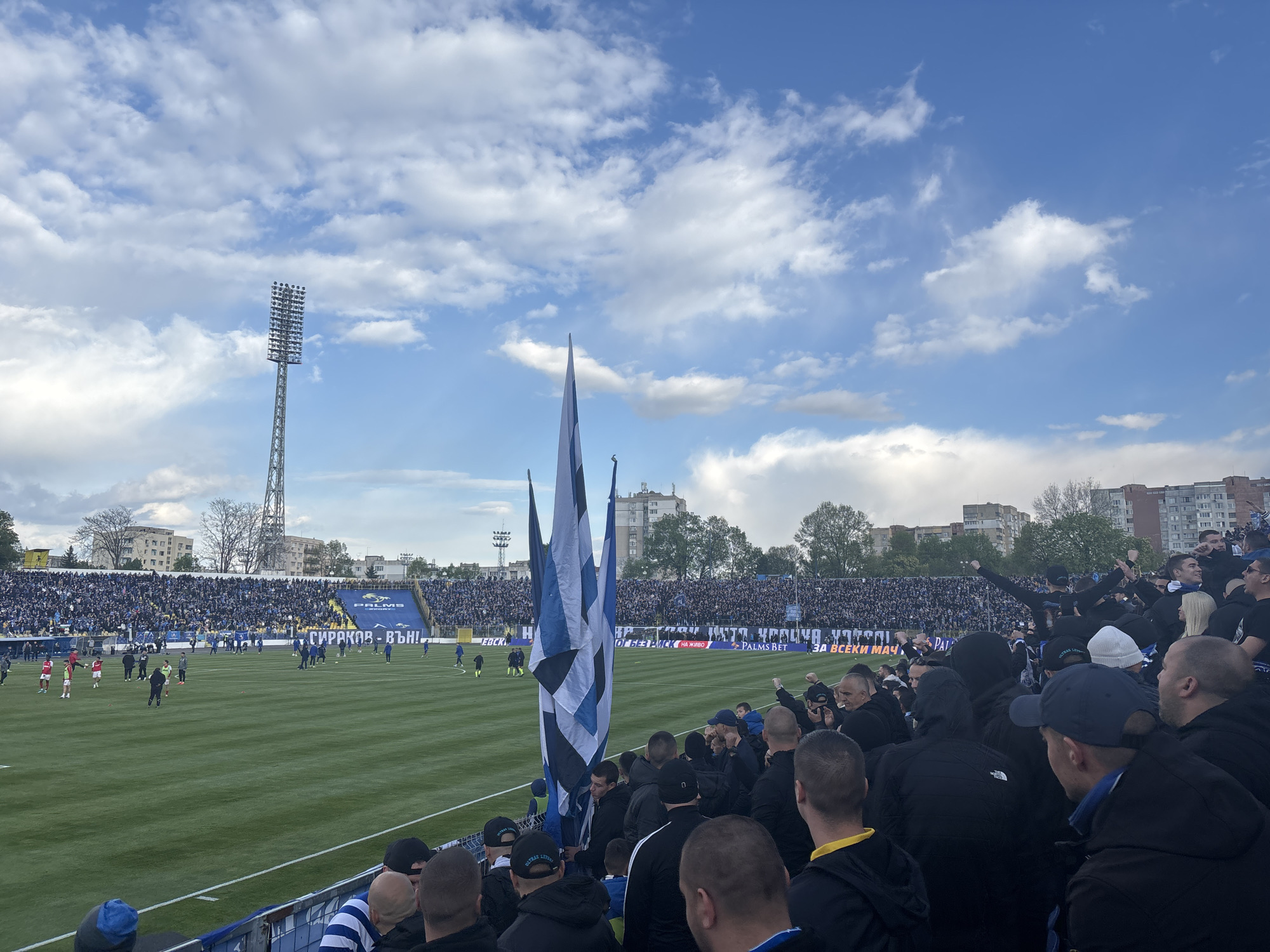 The image shows a stadium filled with people, presumably fans, attending a soccer game. The stadium appears to be the "Slavia" Stadium, located in Sofia, Bulgaria. People are standing and cheering. In the foreground, there are spectators mostly wearing dark clothing. Several are waving large blue and white flags. In the background, there is a soccer field with players, the stands are filled with more spectators. A tall tower with lights stands in the distance. Above, the sky is partially covered with clouds. Advertisements and banners can be seen along the edge of the field.