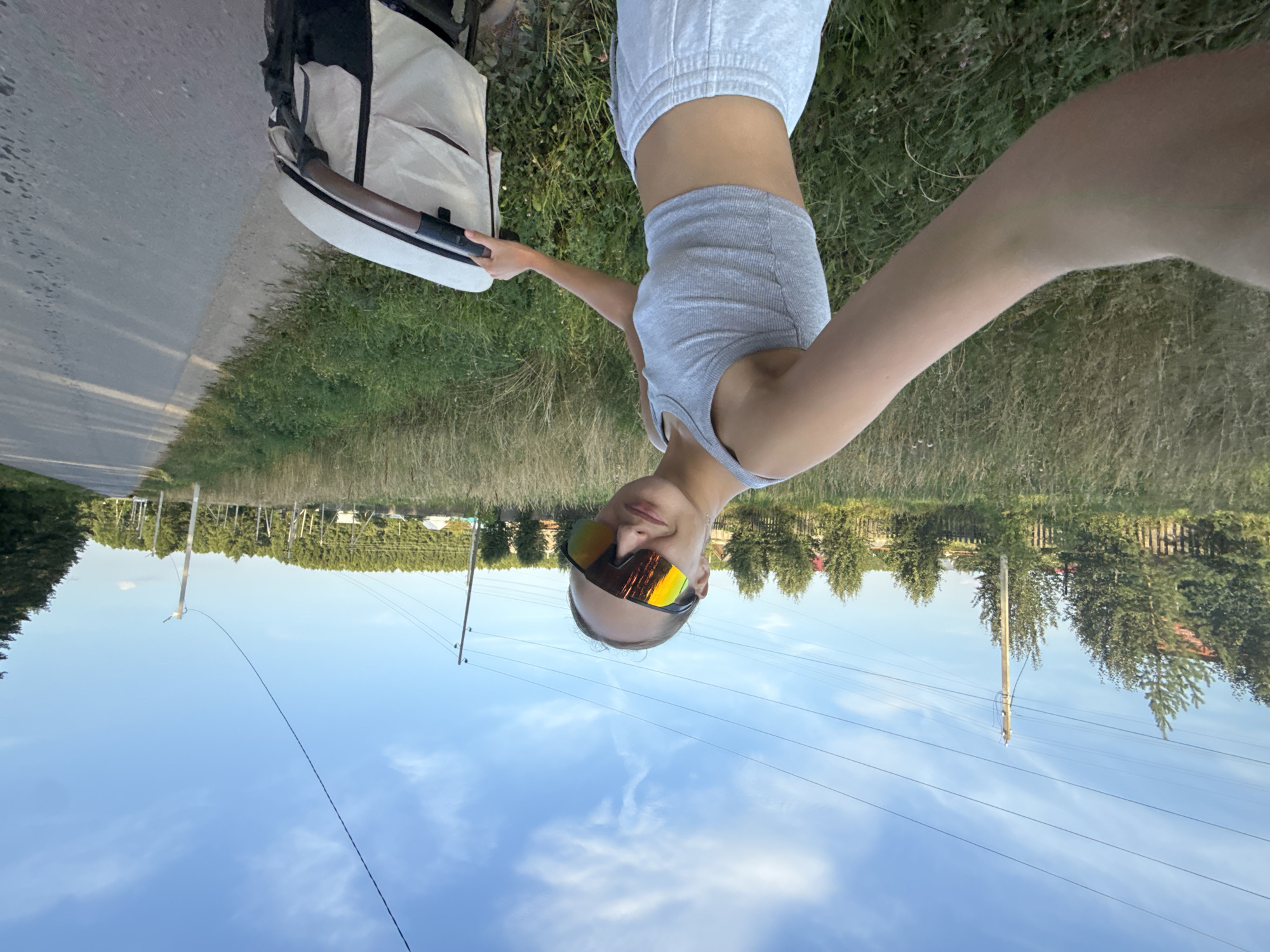 A young woman, wearing a gray tank top and sweatpants, and large, reflective sports sunglasses, is taking a selfie while pushing a beige and black stroller. She is standing on the side of a paved road, with a field of tall, dry grass to her left and a line of trees and houses in the distance. The sky is blue with scattered white clouds. The scene appears to be in a rural area, likely the village of Podshivalovo, Russia, given the presence of wooden fences and power lines often seen in such locations. The woman's expression is a slight smile. The overall lighting suggests it is late afternoon or early evening.