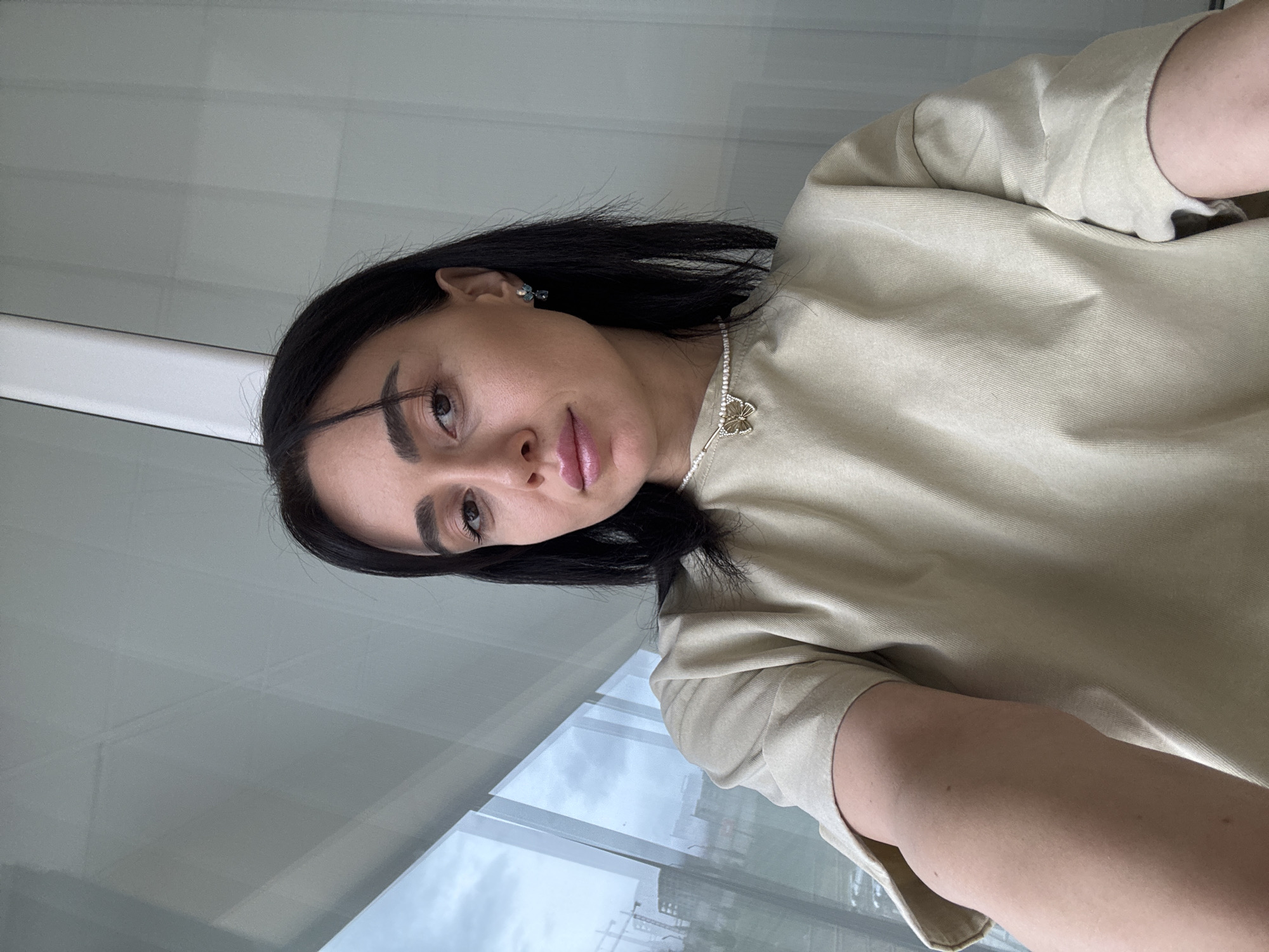 A female individual is centered in a close-up, frontal portrait, looking directly into the camera. She has dark hair, dark eyes, and full lips accented with light pink lipstick; a small mole is present above the right corner of her upper lip. She wears a light beige, possibly ribbed, short-sleeved top, a pearl choker necklace with a gold-toned butterfly pendant, and small dangle earrings featuring blue and white elements in her visible right ear. Her left arm is partially visible in the lower left, indicating a selfie. The background features large, clear windows. Through the windows, an overcast sky and an urban landscape are visible, characterized by multiple construction cranes and partially built structures. Reflections on the window glass show an indoor white ceiling. The scene is situated in the city of Tbilisi, Georgia.