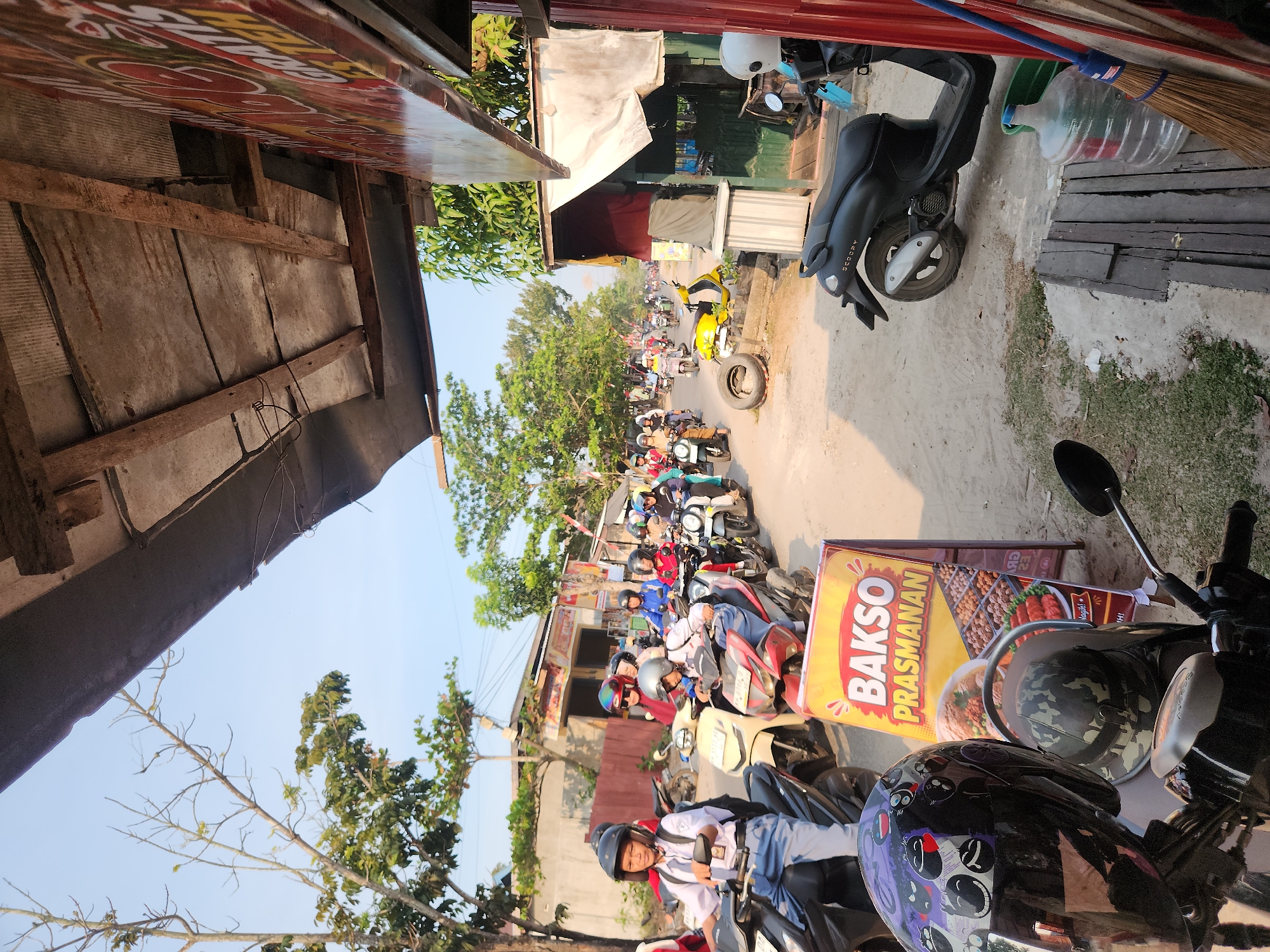 The image captures a street scene in Sampit, Indonesia, likely during daytime. A busy road is filled with numerous motorcycles, many carrying passengers, suggesting a mode of transportation common in the area. Several individuals, including what appear to be students in school uniforms, are riding or waiting on their motorcycles. A prominent A-frame sign advertising "BAKSO PRASMANAN" (a type of Indonesian meatball dish) stands on the roadside, indicating a food vendor or eatery. The environment appears to be a mix of residential or commercial buildings and vegetation. The sky is clear and blue, suggesting fair weather. The foreground shows the handlebars and helmets of parked motorcycles, adding to the sense of a bustling urban environment.