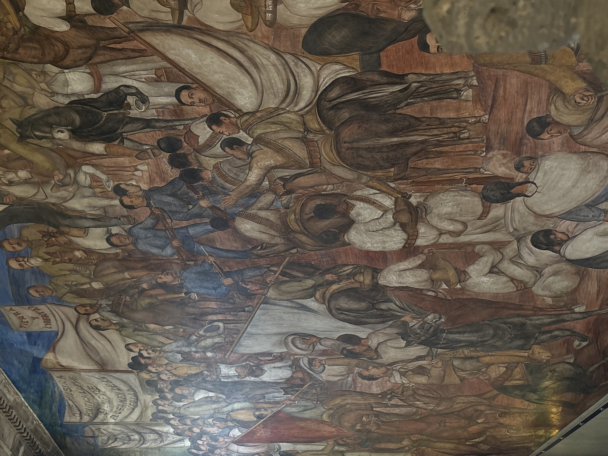 This mural, located in Mexico City, depicts a sprawling and dynamic scene of historical conflict, likely referencing the Mexican Revolution. The artwork is densely packed with numerous figures, both men and women, young and old, engaged in a collective struggle.
**Subjects:** The central subjects are a multitude of people, some on horseback and many on foot. They are dressed in period clothing, with revolutionaries identifiable by their bandoliers and rifles, and others in more civilian attire including sombreros. Several horses are integrated into the composition, some being ridden into battle, while others appear to be fallen or amidst the chaos. Weapons such as rifles and possibly spears are carried by many. Children and women are present, some seemingly supporting the fighters or caught in the struggle.
**Activity or event taking place:** The mural captures a moment of intense battle or a revolutionary march. Figures are shown advancing, fighting, falling, and carrying banners. The overall impression is one of a large-scale popular uprising or a significant conflict.
**Notable details:** The color palette is dominated by earthy tones, browns, and ochres, with splashes of blue in what appears to be a sky or background on the left. Faces express a range of emotions, from grim determination and defiance to pain and sorrow. The style is characteristic of Mexican muralism, known for its epic scale and social realism.
**Visible text:** Two prominent white flags carry inscriptions. On the upper left, the words "TIERRA Y LIBERTAD" (Land and Liberty) are clearly visible, a famous slogan of the Mexican Revolution, particularly associated with Emiliano Zapata. Below it, another flag reads: "NUESTRA POLITICA ES EL GERMEN DE LA VERDAD" (Our politics is the germ of truth).