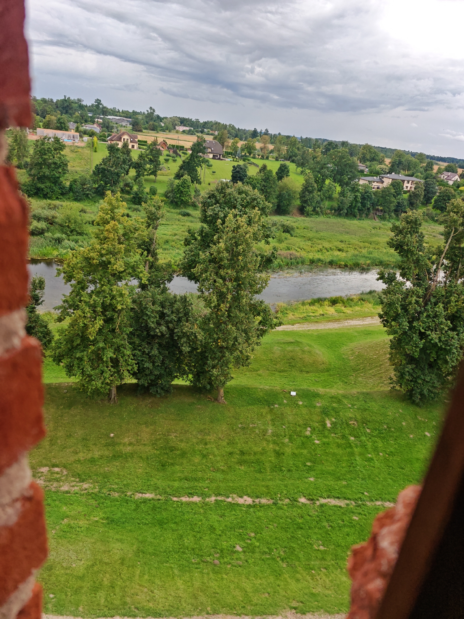 The image captures a view from a brick structure overlooking a river, lush green fields, and a village. The sky is overcast with dramatic clouds. In the foreground, the rough texture of red brickwork is visible, framing the scene. Beyond the immediate grassy area, a winding river flows, bordered by dense green trees and vegetation. Across the river, the landscape rises gently, dotted with houses with beige and red roofs, surrounded by trees. Some houses appear to be modern residences, while others might be older structures. In the distance, more trees and the hazy outline of a larger settlement are visible under the cloudy sky. The overall impression is one of a peaceful, rural setting in a temperate climate. This view is consistent with the area surrounding Bauska Castle in Bauska, Latvia, which overlooks the confluence of the Mūsa and Mēmele rivers, forming the Lielupe.