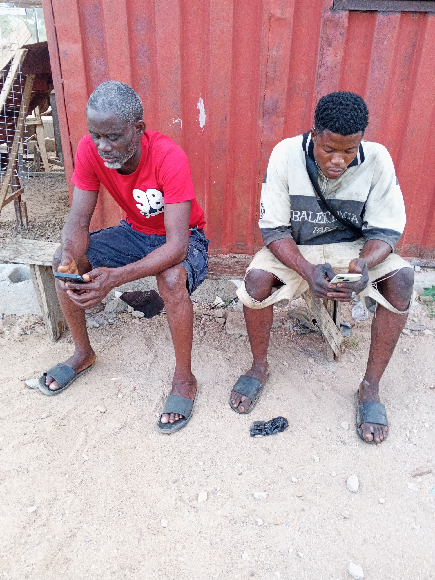 Two men sit side-by-side outdoors in Lokoja, Nigeria. The man on the left, older with gray hair, wears a red t-shirt with the number "98" on it and dark shorts. The man on the right, younger with black hair, wears a white and gray Balenciaga top and light-colored shorts. Both men are looking at and using smartphones. They are sitting on separate wooden stools, placed on a sandy ground. Behind them is a red corrugated metal wall, partially visible. In the background to the left, there is a wooden structure possibly related to a goat pen. Between the men's feet is a small piece of dark trash on the ground. They are both wearing rubber sandals.