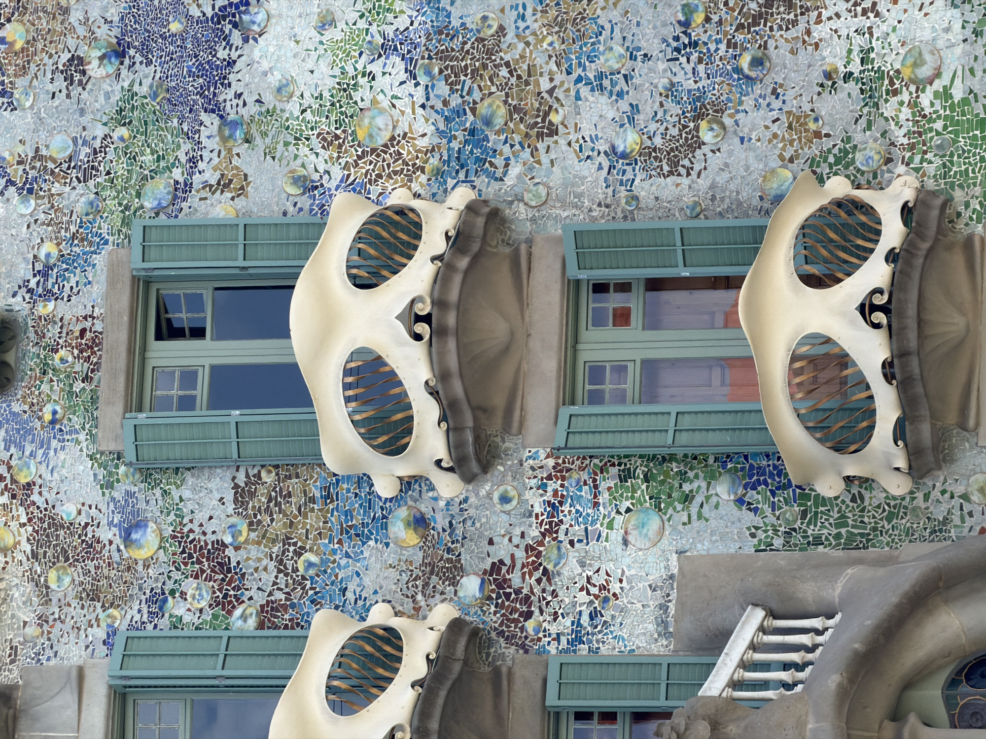 The image displays a close-up view of the facade of Casa Batlló, a famous building in Barcelona, Spain, designed by Antoni Gaudí. The facade is covered in a mosaic of colorful tiles, primarily in shades of blue, green, and brown, with circular ceramic elements that resemble bubbles. Several windows with green shutters are visible, as are two distinctive balconies with bone-like structures and wrought-iron railings. The architecture is organic and fluid, characteristic of Gaudí's modernist style. The lighting suggests it is daytime, with soft shadows indicating an overcast sky or indirect sunlight. There are no people or animals visible in the frame. The overall impression is one of artistic and architectural detail.