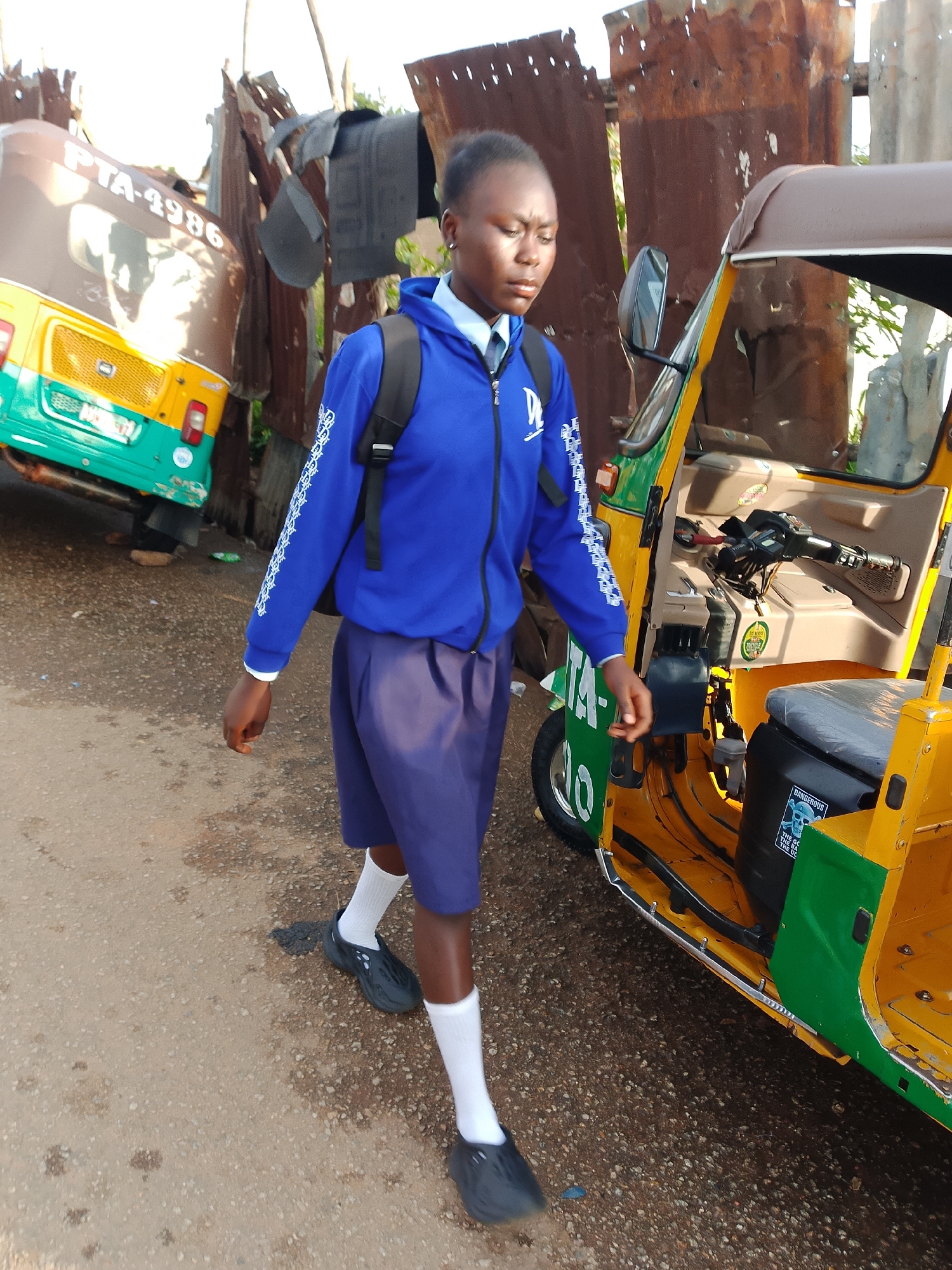 A young person, possibly a student, walks beside a yellow and green auto-rickshaw. The person is wearing a blue jacket with white trim on the sleeves and collar, a dark blue skirt, white socks, and dark shoes. They have a black backpack on. The auto-rickshaw appears to be parked on a dirt or gravel road. In the background, there is a rusty corrugated metal fence and another auto-rickshaw, partially visible, with the license plate "PTA-4986". The scene appears to be outdoors in an urban or semi-urban environment. The lighting suggests it is daytime.