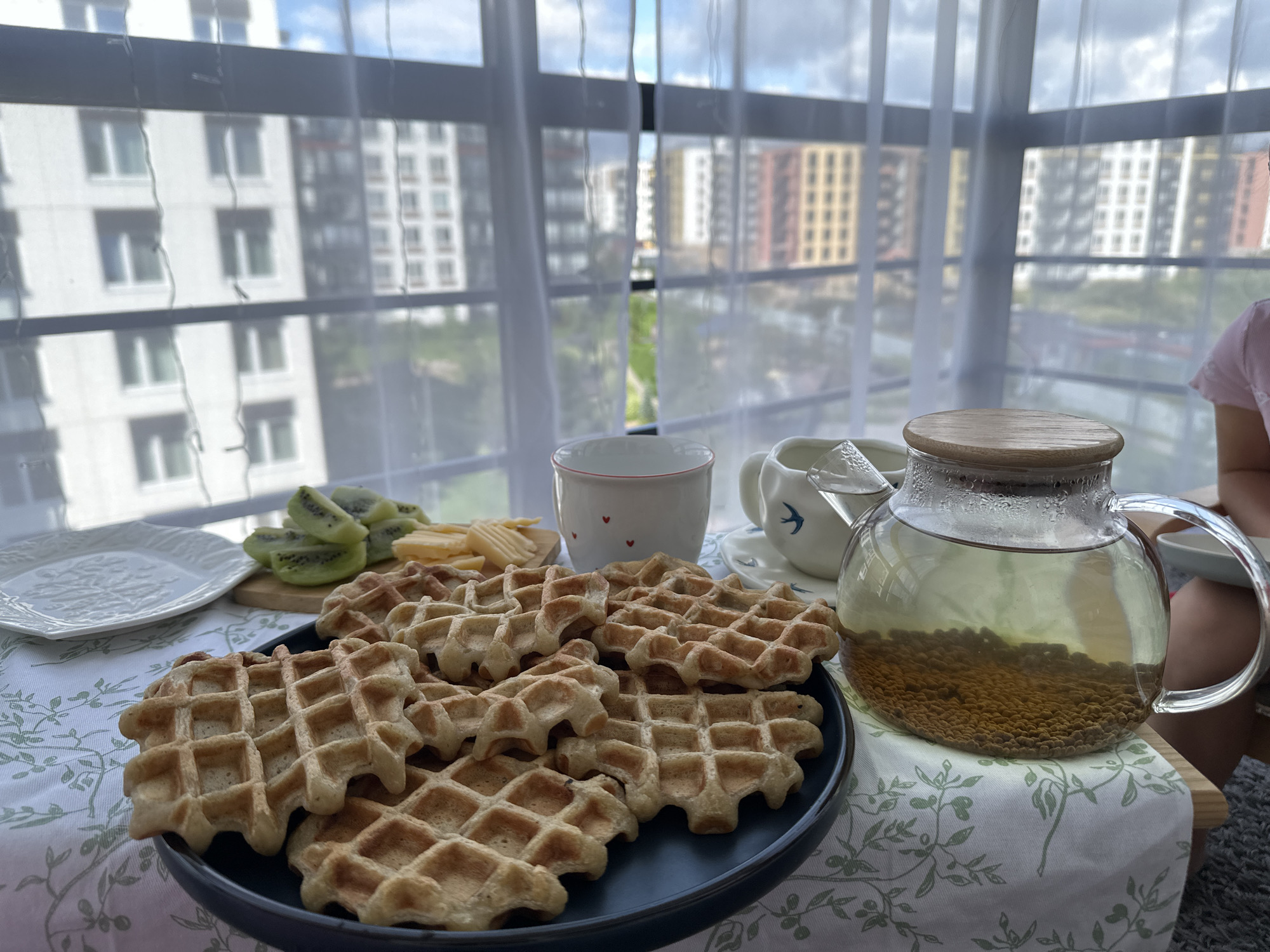 An outdoor breakfast table is set on a balcony overlooking an urban landscape. The scene features a plate of waffles, sliced kiwi, cheese, a cup, a creamer, and a teapot filled with herbal tea. The background shows apartment buildings through sheer curtains, with a clear sky visible above. The image is a still life, with no discernible action or interaction. The setting appears to be Vsevolozhsk, Russia, based on the architectural style of the buildings.