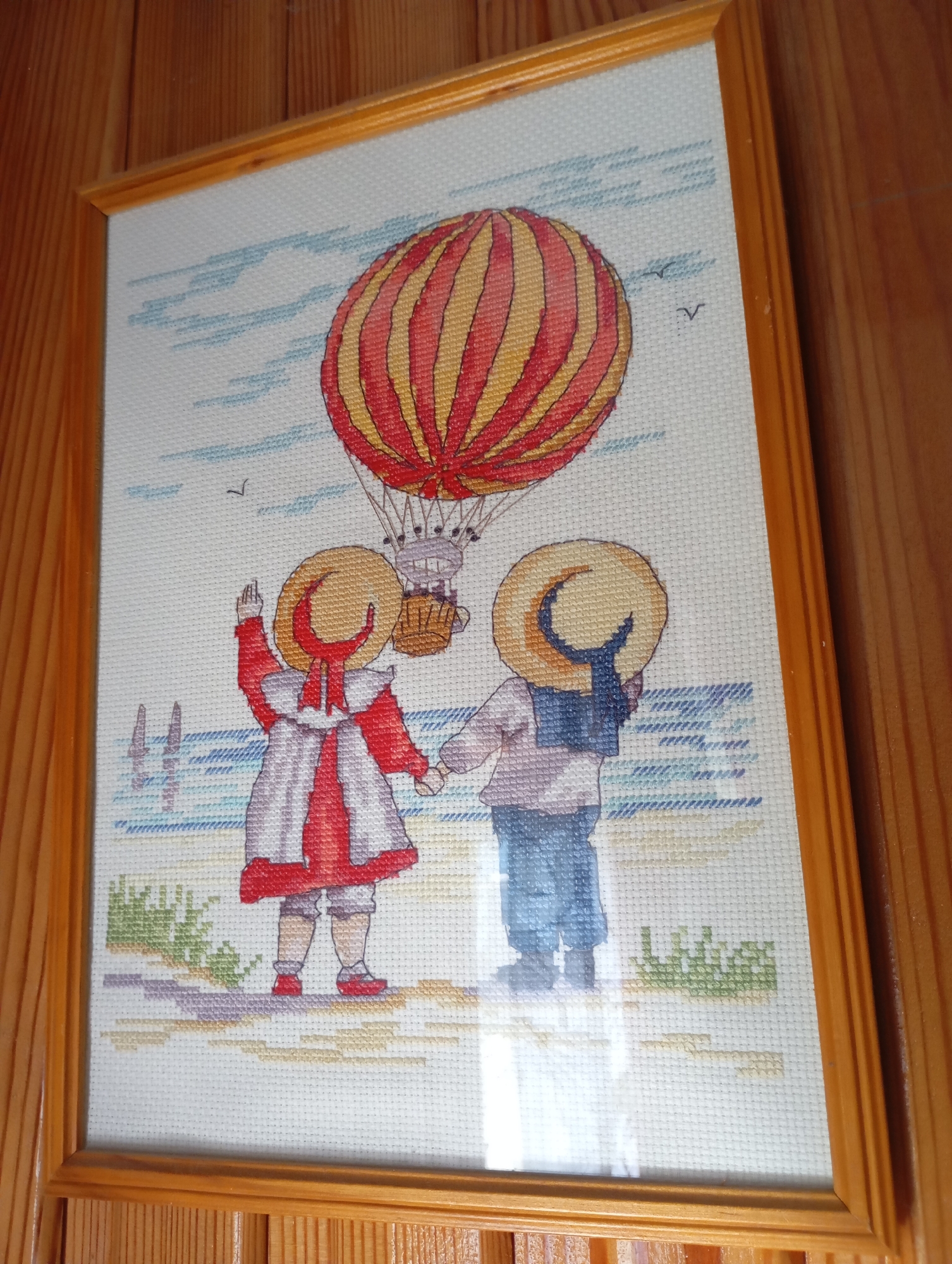 The image shows a framed cross-stitch artwork depicting a hot air balloon over a beach scene. Two children, one in a red and white dress and the other in blue overalls and a sailor collar, stand holding hands on the beach. The hot air balloon is orange and yellow striped, with a basket hanging below. The background shows a blue and white sky with a few small birds and faint clouds. The beach has green grass and a sandy shore with blue water. The artwork is framed in a simple wooden frame and hangs on a wooden wall. The location of Льниха, Russia is not depicted in this image.