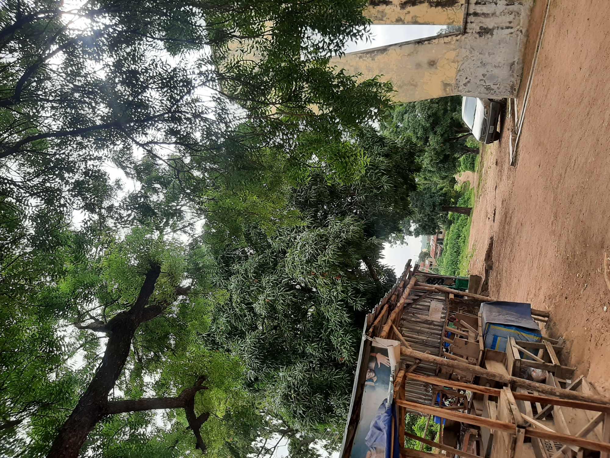 The image depicts a dirt road in Ogbomosho, Nigeria, with a small wooden market stall on the left and a light-colored car parked on the right. The market stall is rustic, constructed from wood and corrugated metal, and appears to be selling various items, though they are not clearly identifiable. A poster of two men is affixed to the side of the stall. In the background, there are several trees with dense green foliage, some houses, and lush vegetation. A large, weathered concrete structure, possibly an archway or part of a building, stands to the right of the car. The sky is overcast.