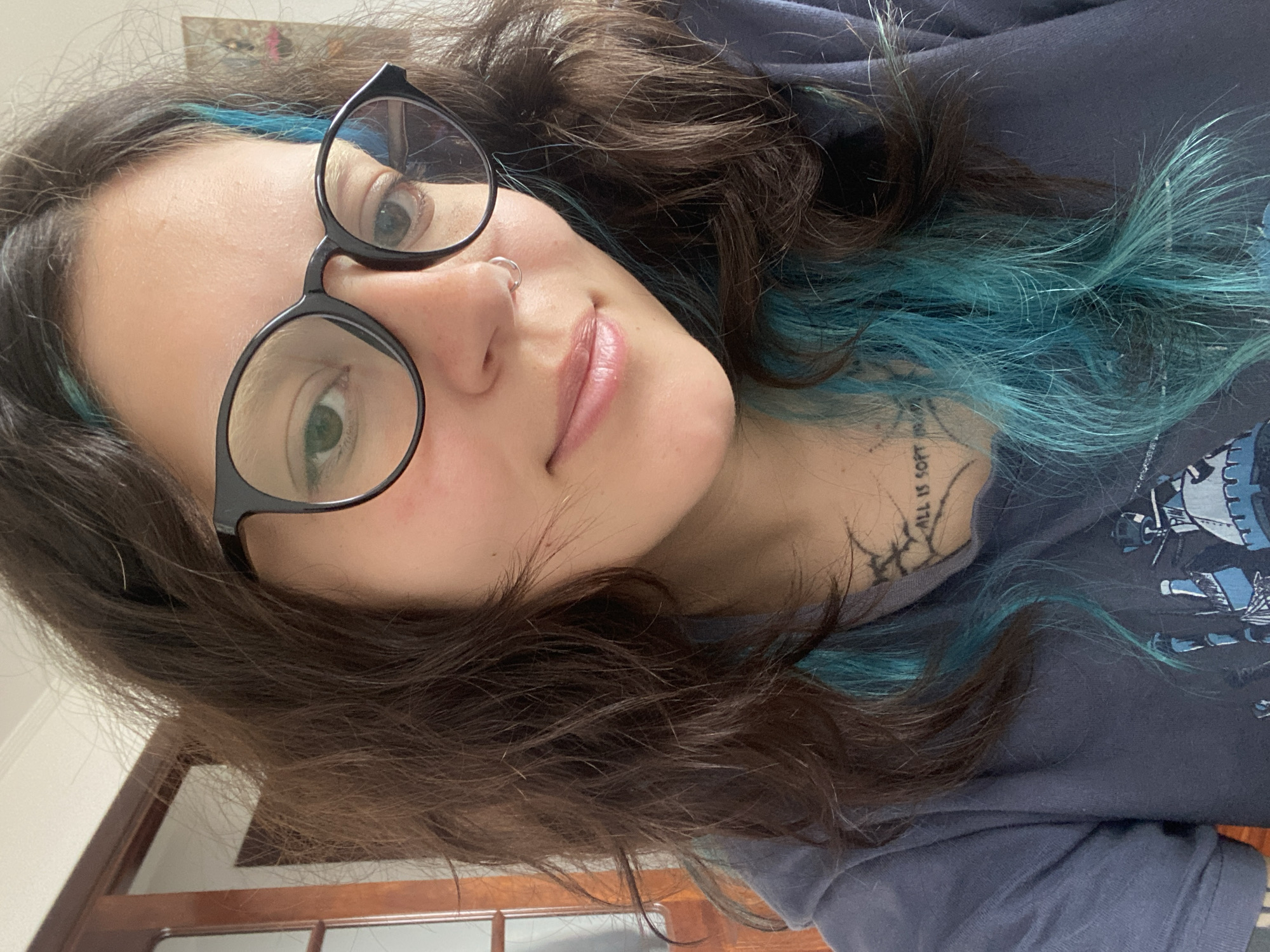 A woman with brown hair and blue streaks, wearing round glasses and a nose ring, looks at the camera. She is wearing a dark blue t-shirt with a design on it and has a tattoo on her neck. A partially visible wooden door is in the background. The location is unknown.