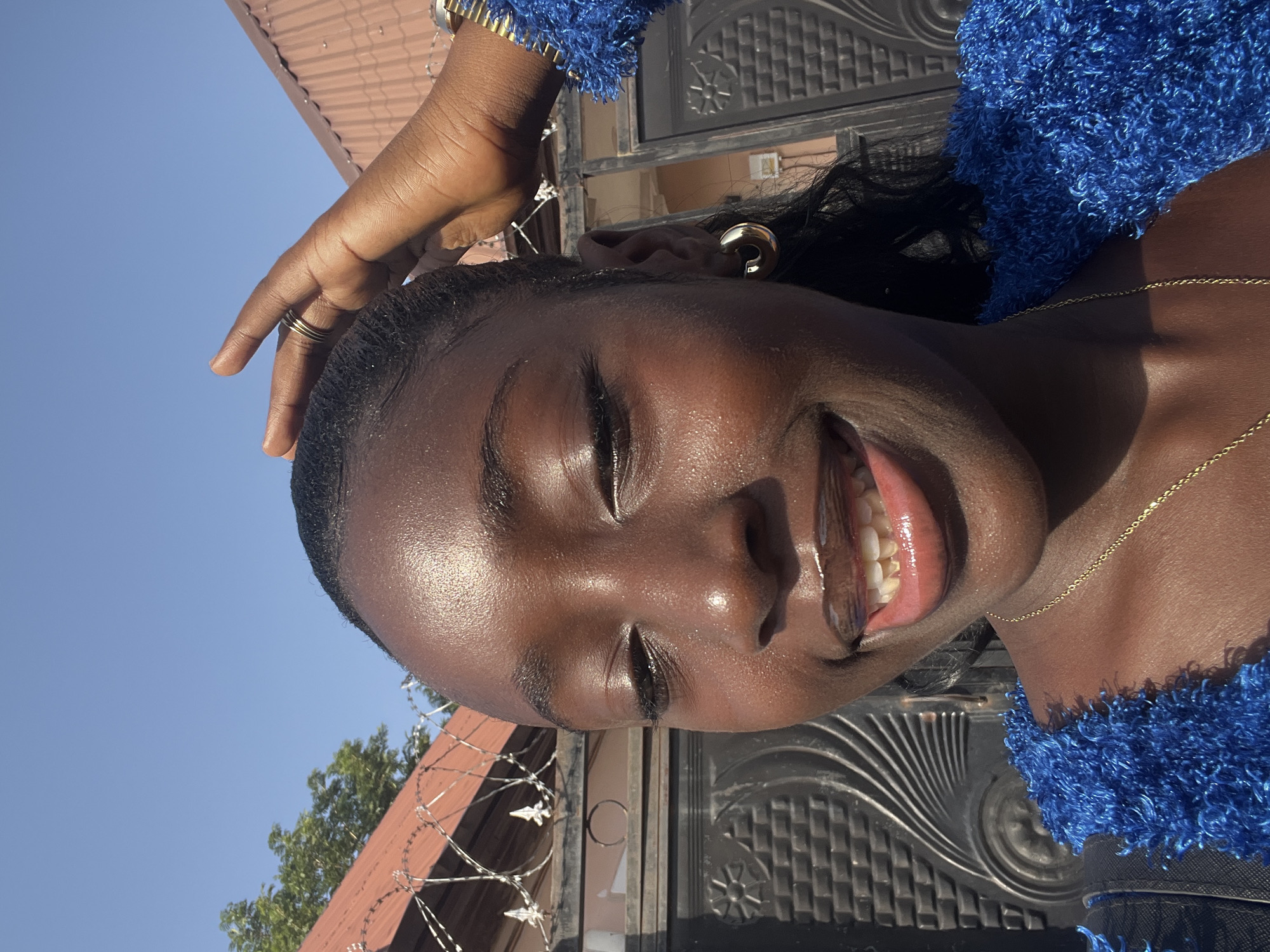 A close-up shot looking up at a woman of dark complexion, smiling with her eyes closed. Her right hand is raised to her forehead. She is wearing a blue, textured top, a gold necklace, and gold earrings. Her teeth are visible. Behind her is a wrought-iron gate, a brown roof with barbed wire, and a clear blue sky. The location appears to be in a sunny outdoor area in Abuja, Nigeria.