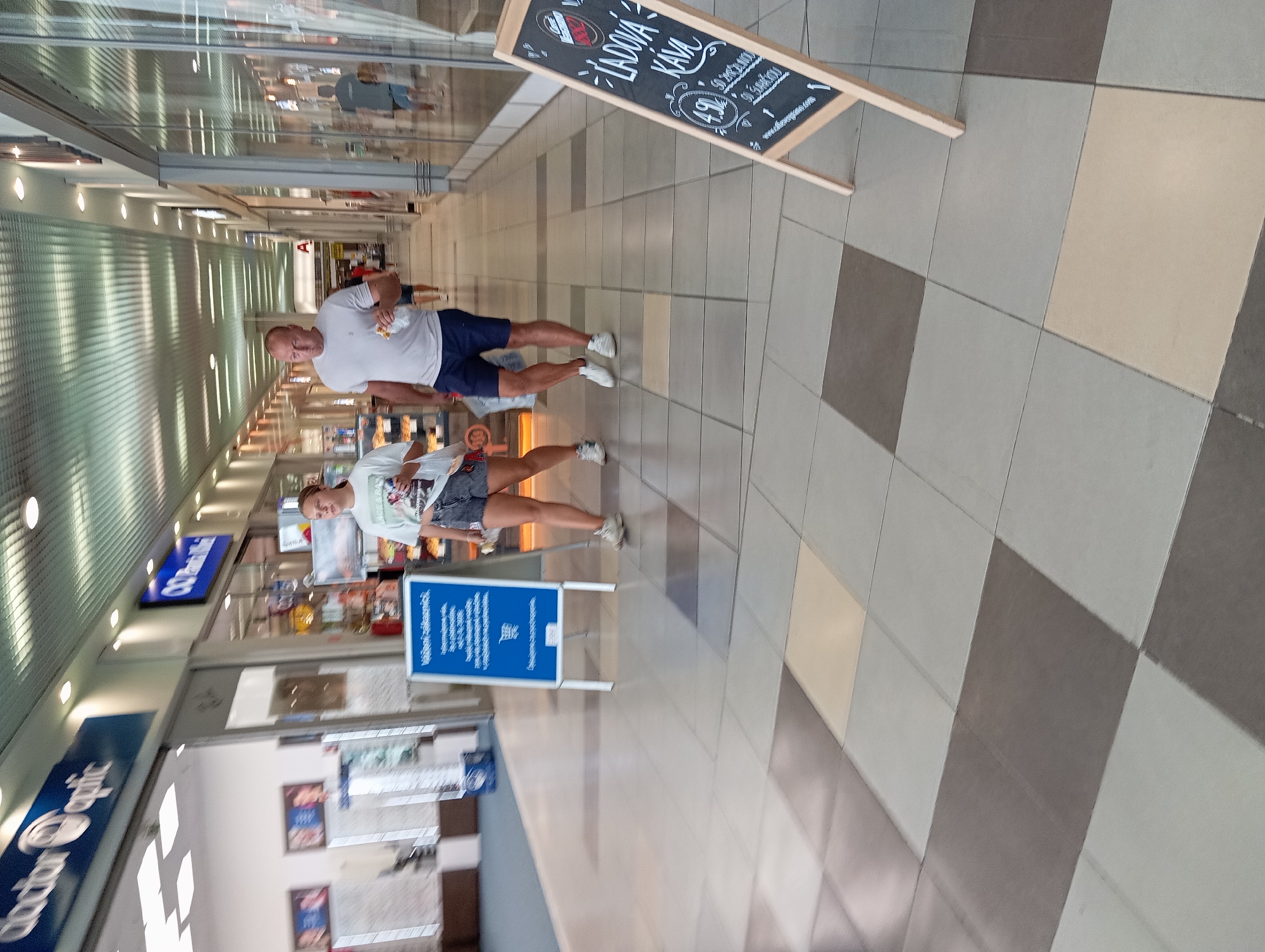 The image shows an interior of a shopping mall in Nitra, Slovakia. Two people are walking down the hallway. On the left, a young woman in a white t-shirt and denim shorts walks towards the camera, holding a white bag. To her right, a man in a white t-shirt and blue shorts walks away from the camera, holding a white sandwich. In the foreground on the right, there is a chalkboard sign advertising "LÁDOVÁ KÁVA" (iced coffee). The hallway has a tiled floor with a checkered pattern of gray and beige tiles. Fluorescent lights are visible on the ceiling. Stores are visible on either side of the hallway.