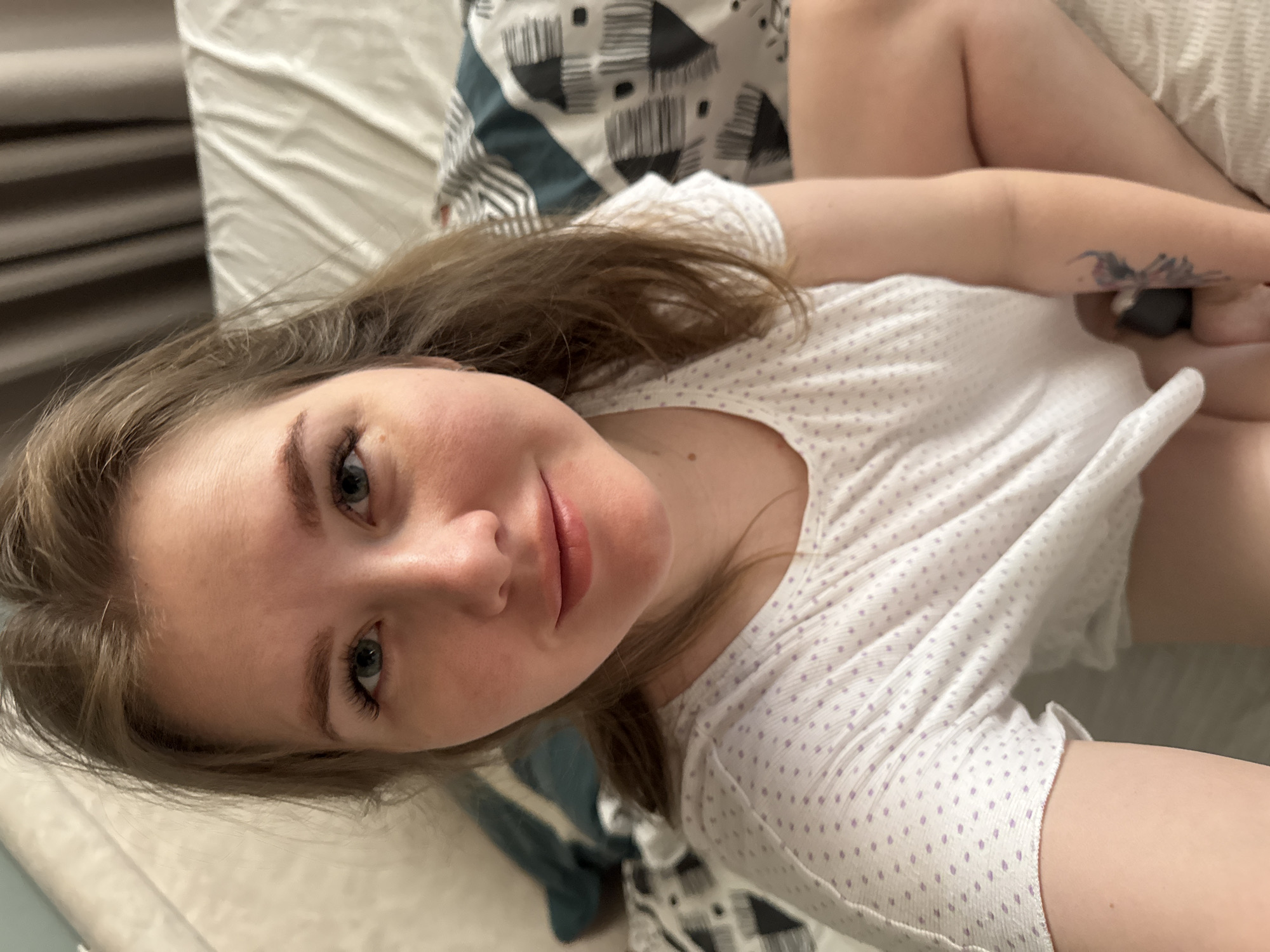 The image depicts a young adult female subject in an indoor setting. The specified location of Gornyi (Dvurechenskoe S/P), Russia, cannot be corroborated by visual evidence within the image itself.
The female, with light skin, light brown hair, and blue-green eyes, is positioned facing directly towards the camera, suggesting a selfie. She exhibits a slight, closed-mouth smile. She wears a white, short-sleeved top patterned with small purple polka dots. A multi-colored tattoo, appearing to depict a butterfly or similar insect, is present on her right forearm. Her left hand is positioned near her lap, holding a small, dark, unidentifiable object. Her lower body is partially visible, showing bare skin on her thighs.
The background indicates an interior space, likely a bedroom. A patterned duvet cover featuring black, white, and teal geometric designs is visible behind the subject and to her right. A plain light-colored sheet or pillow is situated to her left. Blurred light-colored fabric, possibly a headboard or wall, is present behind her left shoulder, and indistinct dark vertical elements, consistent with curtains, are visible in the upper right background. The lighting suggests an artificial indoor source.