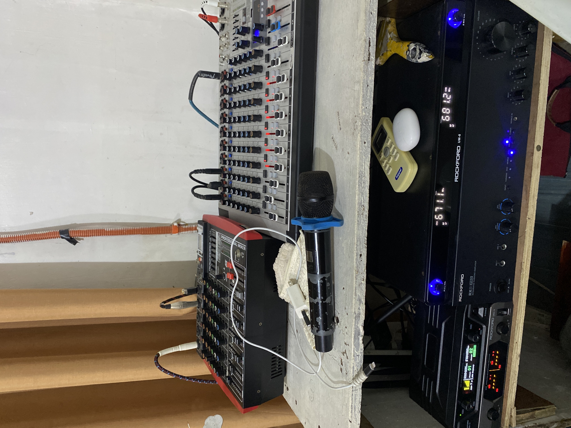 The image depicts an indoor audio equipment setup, potentially located within a residence or small venue in Davao City, Philippines. No individuals are visible in the frame. The scene is dominated by two levels of audio equipment resting on worn, light-colored wooden surfaces. On the upper shelf, from left to right: * A black and red "ROCKFORD RM-7A" professional audio mixer is positioned on the left, featuring multiple input channels, various colored control knobs (black, blue, green, red), and white faders. Several cables, including a patterned blue and white cable, are connected to its inputs. * In the center foreground, a black-bodied dynamic microphone with a dark blue grille is lying horizontally. The microphone exhibits significant paint wear on its body. It is partially wrapped in a small, off-white, textured cloth. A white USB-A to Lightning cable is connected to the microphone, with the USB-A end plugged into a small white power adapter cube resting on the cloth. * To the right, a silver-grey and black professional audio mixer with numerous input channels, black and blue control knobs, and white faders is present. Several red LEDs are illuminated on some of the faders. Cables are connected, including a black XLR cable, a blue cable with "Fender" branding, and red and white RCA cables. The background consists of a light brown pleated curtain on the left and a white, textured wall on the right. A vertical orange corrugated conduit pipe, secured to the wall with black tape, runs along the right side of the frame. On the lower shelf, from left to right: * A black "ROCKFORD URM-T" UHF Wireless Microphone Receiver is positioned on the left. Its digital display shows frequency information (e.g., "650.00-660.00 MHz") and channel indicators ("01," "02"), along with signal strength bars. Several orange-red LEDs are illuminated below the display. * Next to it, a black "ROCKFORD RX8 PRO" Integrated Karaoke Amplifier is displayed. Its digital readout shows "671.1 MHz" and "681.2 MHz." A prominent blue illuminated knob is visible on its right side. Below the display, various controls include a "MIC ECHO" knob, input selectors ("INPUT," "KARAOKE," "DVD," "BLUETOOTH," "S-BASS," "MOBILE IN"), and knobs for "BASS," "MID," "TREBLE," and "VOLUME." * Resting on top of the "ROCKFORD RX8 PRO" amplifier are three items: a yellow/beige remote control with "joppel" branding on the left, a small white oval object (possibly a wireless earbud case) in the center,