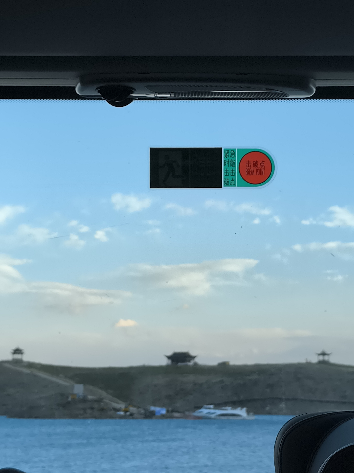 An image taken from inside a vehicle, looking through a window at a bright blue sky with scattered clouds. In the foreground, there is a close-up view of the vehicle's interior, including a black ceiling-mounted component with a small camera. A sticker is visible on the windshield, featuring a running person icon and text in both Chinese and English indicating "Emergency Break Point."
The background shows a landscape across a body of water. On the far shore, there is a sloped green hill dotted with construction equipment and what appears to be scaffolding. Further back on the hill are several traditional Chinese-style structures with distinctive roofs. A white high-speed boat is navigating the blue water. The overall scene suggests a bright, possibly daytime setting, with a mix of urban development and natural scenery. The location is identified as Unknown, China.