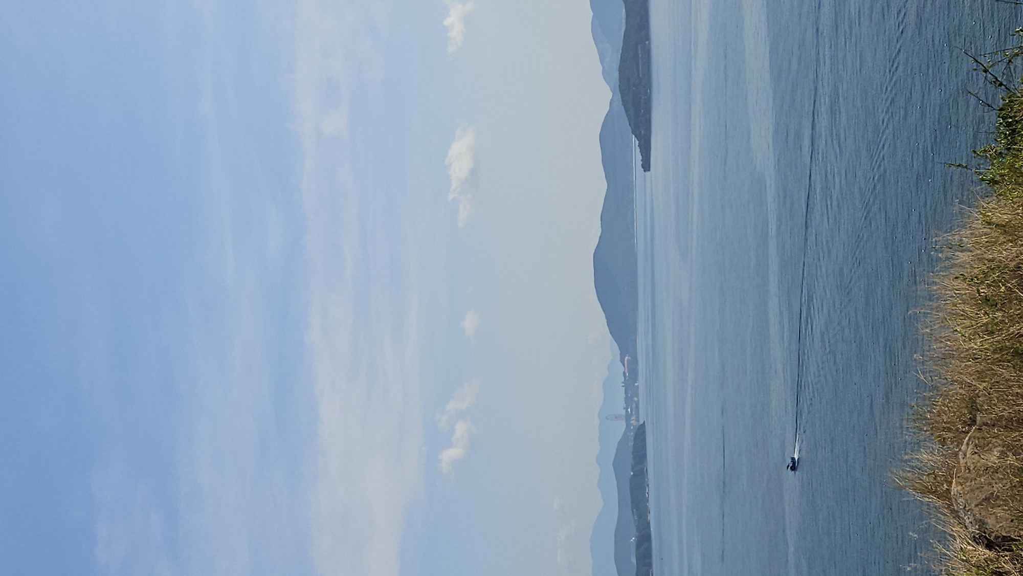 The image depicts a coastal view in the city of Nam Natural Heritage Area Trang, Vietnam. In the foreground is a grassy shoreline with green vegetation. The middle ground features a body of water with a boat moving across its surface, leaving a wake. In the background, there are mountain ranges silhouetted against a light blue sky dotted with a few clouds.