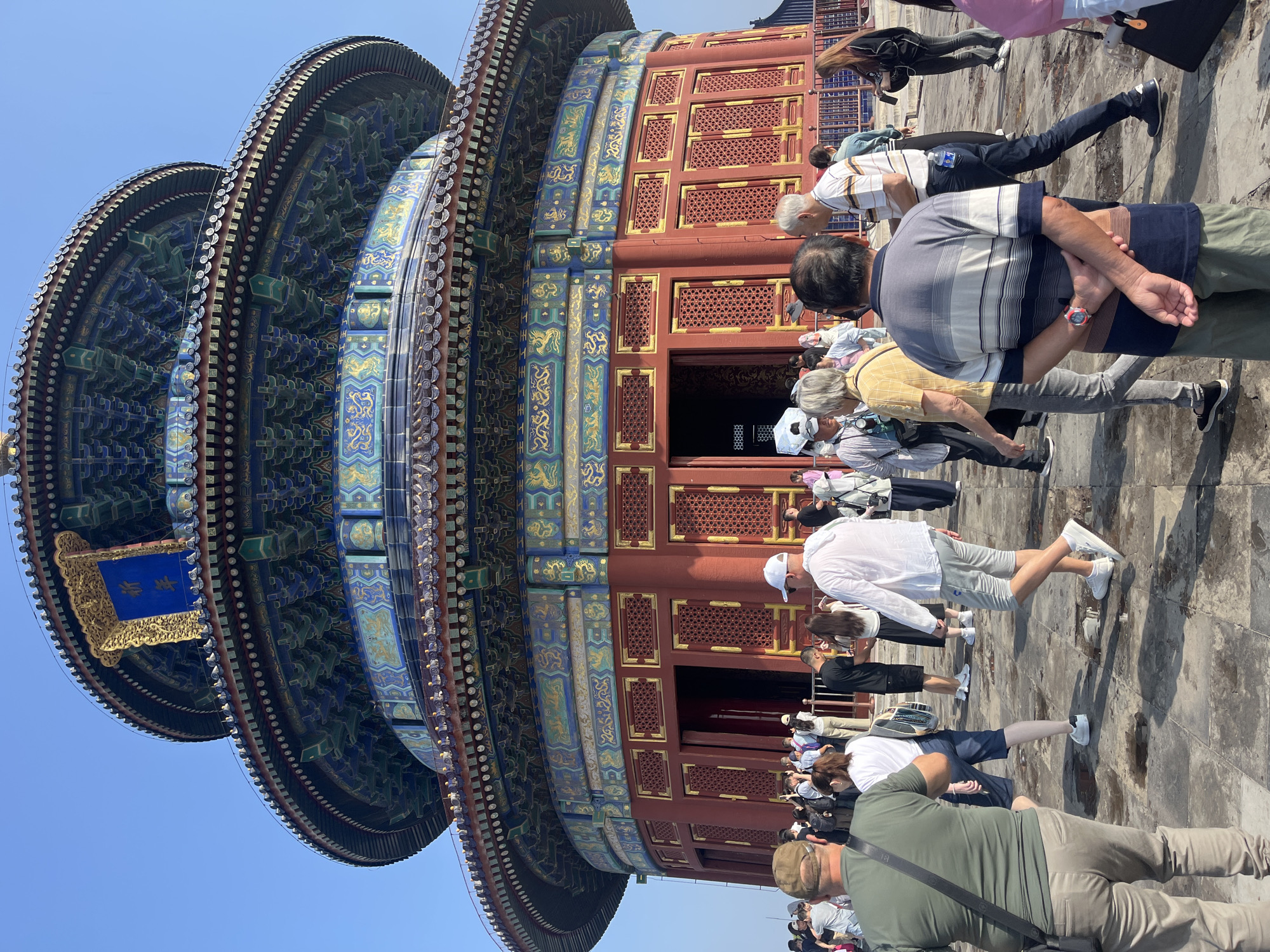 The image shows the Hall of Prayer for Good Harvests at the Temple of Heaven in Beijing, China. The structure is a large, circular, three-tiered building with a blue-tiled roof and red walls. The roof is adorned with intricate carvings and paintings in shades of blue, green, and gold. The walls are decorated with golden latticework and decorative panels.
A crowd of tourists is gathered in front of the building, some walking and others looking at the architecture. They are dressed in casual clothing, suggesting a warm day. The ground in front of the temple is paved with stone, and there are some puddles of water on the ground, indicating recent rain or cleaning. The sky is clear and blue. The scene captures a moment of cultural tourism, with visitors admiring a significant historical landmark.
