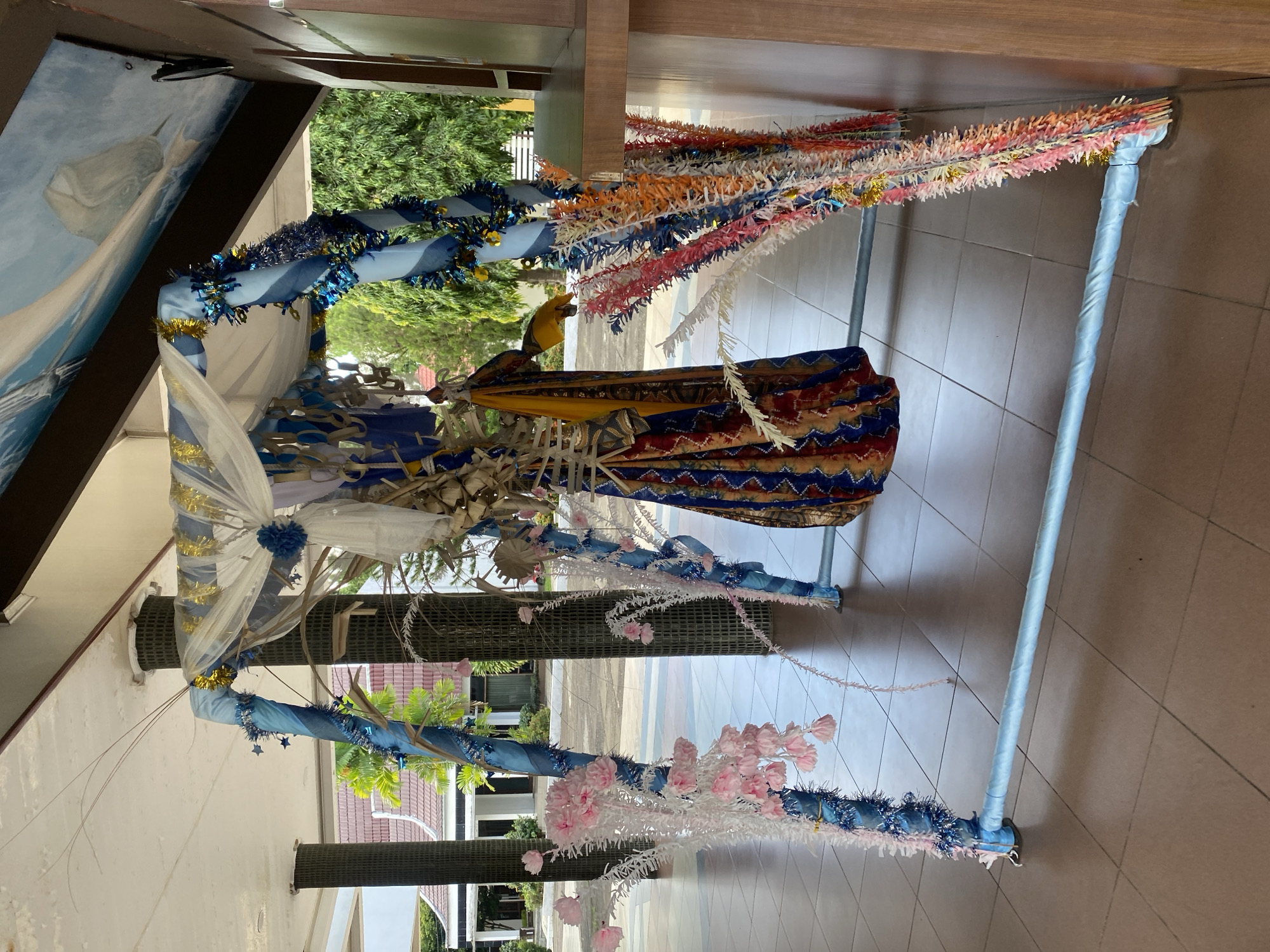 The image shows an outdoor pavilion decorated with blue and white fabric, tinsel, and various floral and fringed ornaments. The pavilion has a square frame constructed from metal poles, with diagonal supports and a canopy. The poles are wrapped in blue fabric, and adorned with blue and gold tinsel, as well as blue stars. Hanging from the canopy are white and sheer fabrics, and a colorful, patterned fabric is draped in the center. On the left side of the pavilion, white branches with pink floral accents are attached to the poles. On the right side, bundles of colorful fringed decorations hang down. In the background, there are trees, a building with a red roof, and a paved area. The flooring of the pavilion is tiled. A painting of a whale is visible on the ceiling of the pavilion. The overall impression is of a festive or ceremonial decoration. The location is identified as a pavilion in Banjarbaru, Indonesia.