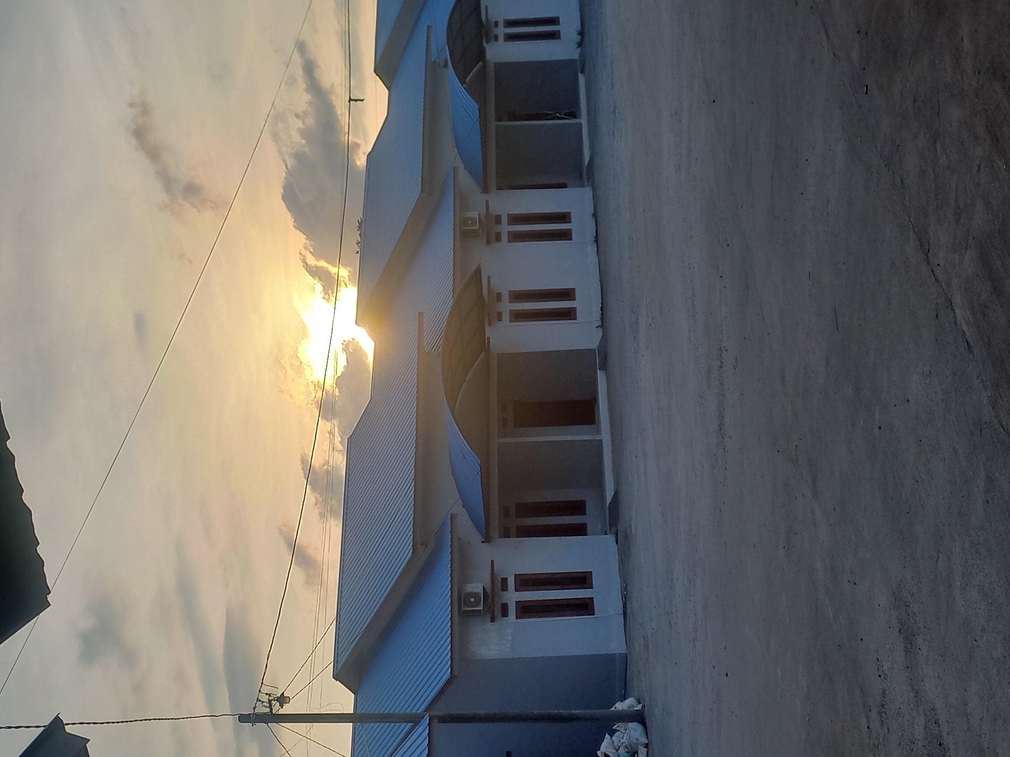The image captures a building in Puloblang, Indonesia, bathed in the light of the setting sun. The building is multi-storied, with a pale blue roof and white walls. Its architecture is characterized by a series of identical bays or sections, each featuring a covered outdoor area with pillars. Each section also contains at least one window, noticeable by its dark horizontal bars. An air conditioning unit is visible on the side of one section.
The sun is positioned directly behind the building, creating a halo effect and obscuring details. The sky is filled with clouds illuminated by the sunlight, creating a golden hue.
A road or paved area stretches out from the building towards the viewer. Debris, including plastic bags, is visible at the base of the building. Several utility poles and wires are visible in the sky.