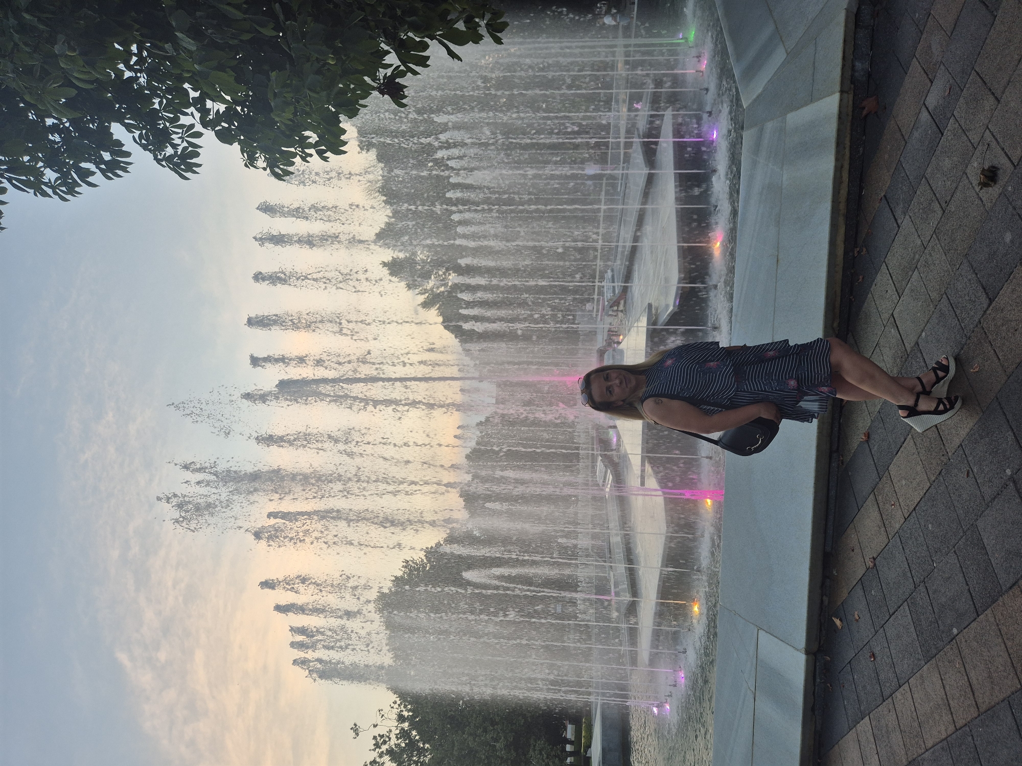 A woman stands in front of a large, illuminated fountain in an outdoor urban setting. The woman, wearing a striped dress and platform sandals, is looking at the camera and has a black purse slung over her shoulder. The fountain displays multiple jets of water, illuminated by colored lights in pink, yellow, and green. Trees are visible in the background, and the sky suggests it is either dawn or dusk. The ground is paved with interlocking stones.